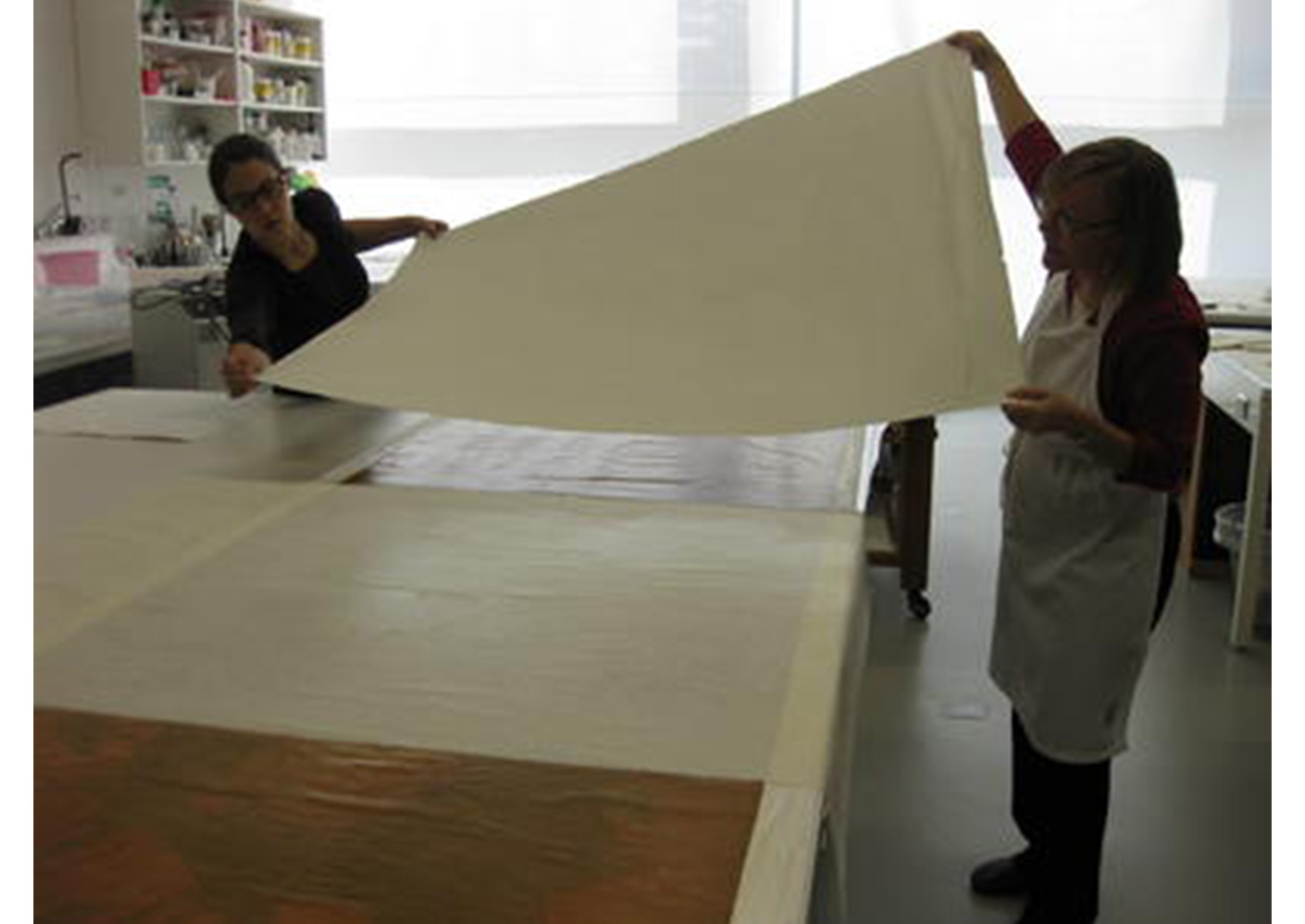 two woman hold a large sheet of paper over a white table with a poster spread across it