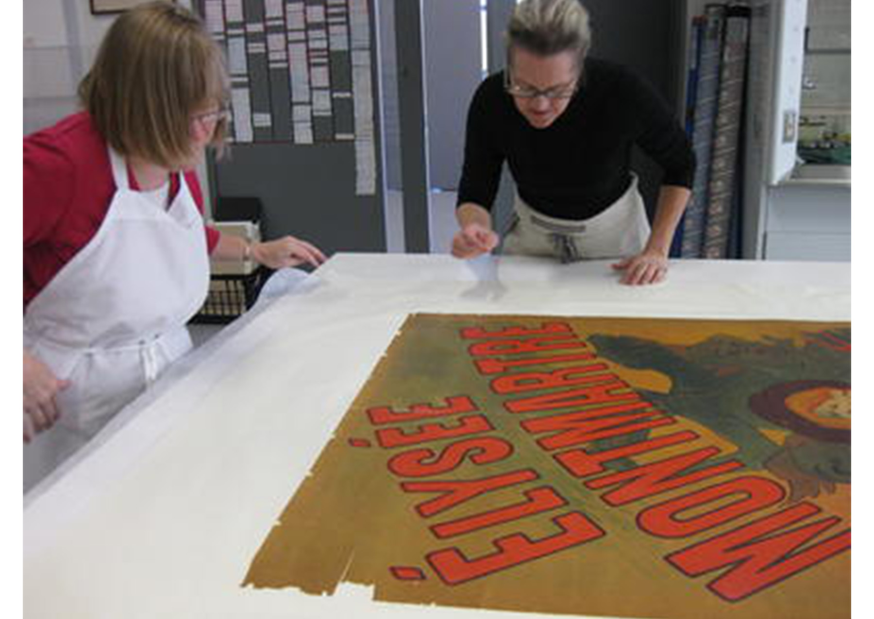 two woman stand over a table, examining the edges of a poster