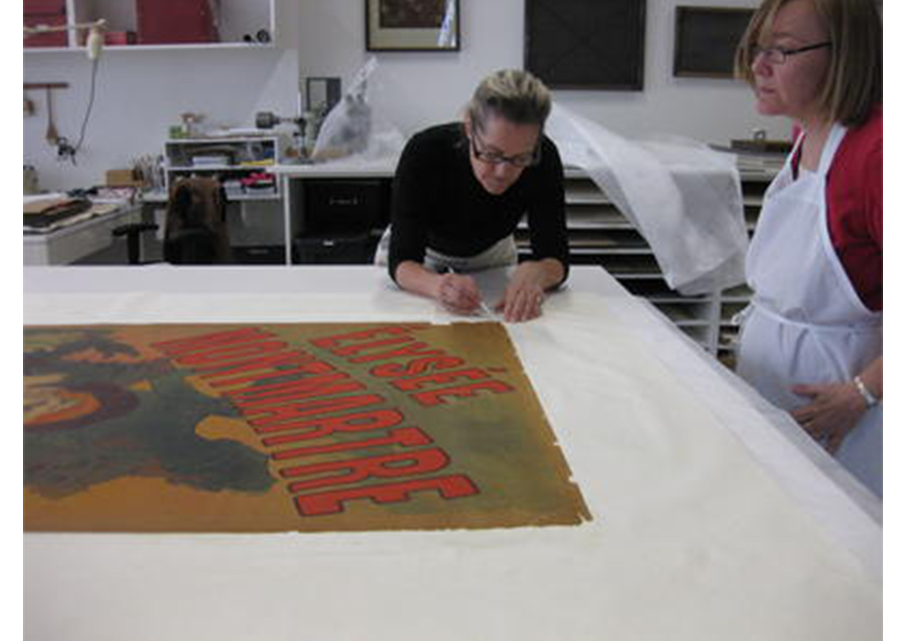 large white table with a poster spread across it; woman tends to a corner of the poster with tweezers; another woman stands over the table looking at the poster