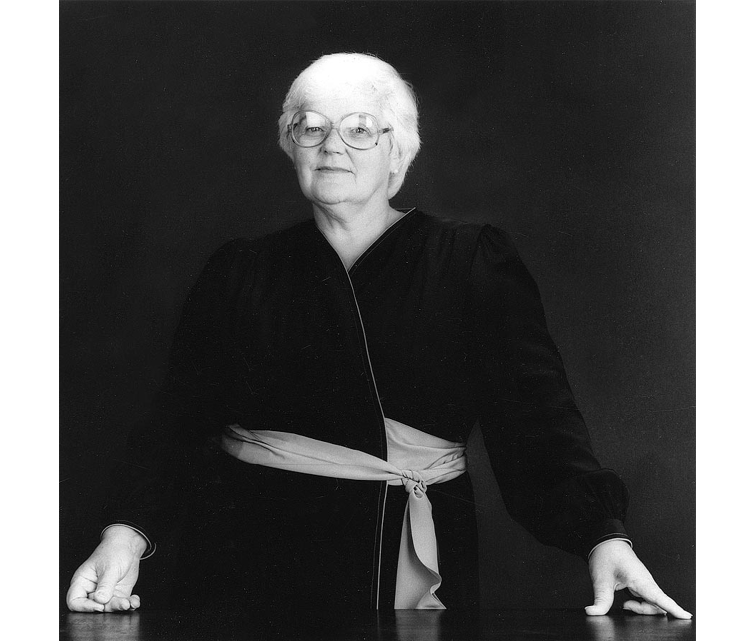 middle aged white haired woman standing behind a table with both hands on the table, plain dark backdrop behind