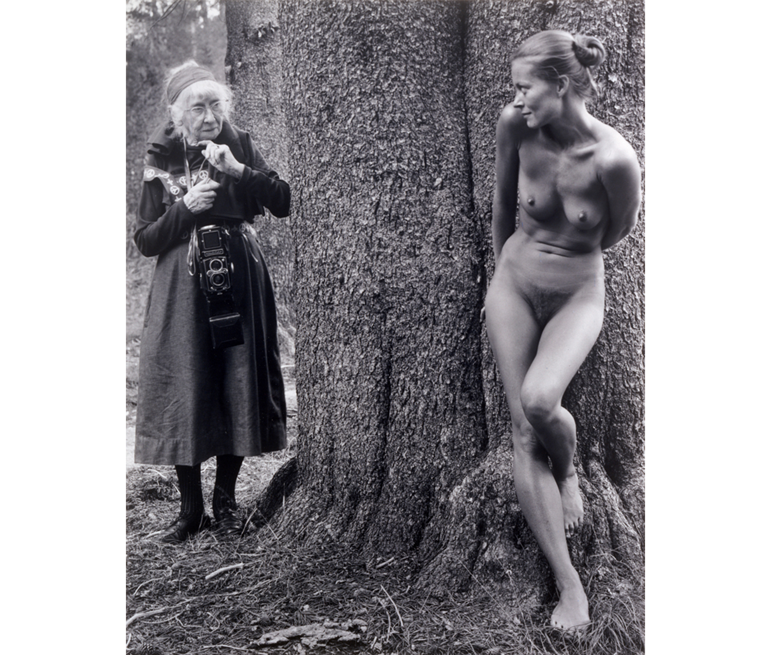 older dressed woman to the right of tree looking at nude woman to the left of tree