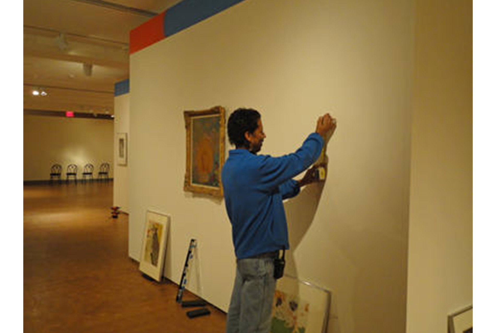 man stands in an art gallery, holding a measuring tape to the wall