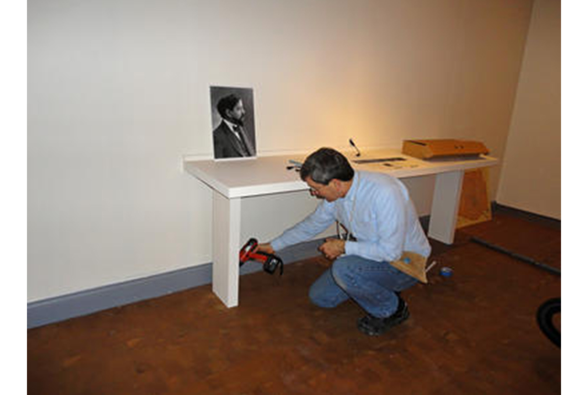 man sits in an art gallery below a table holding a power drill; photograph of profile view of man sits on the table above him