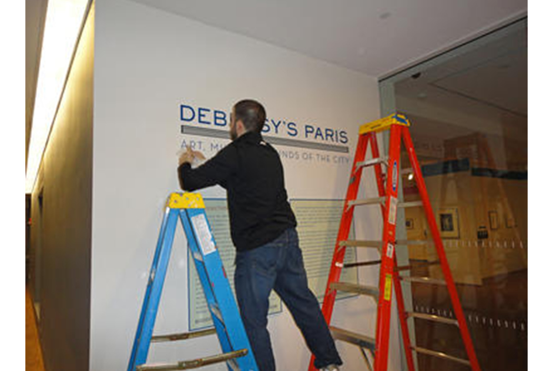 man stands on two ladders, one blue and one orange, installing text onto a wall display. text reads, "DEBUSSY'S PARIS: ART, MUSIC, AND THE SOUNDS OF THE CITY"