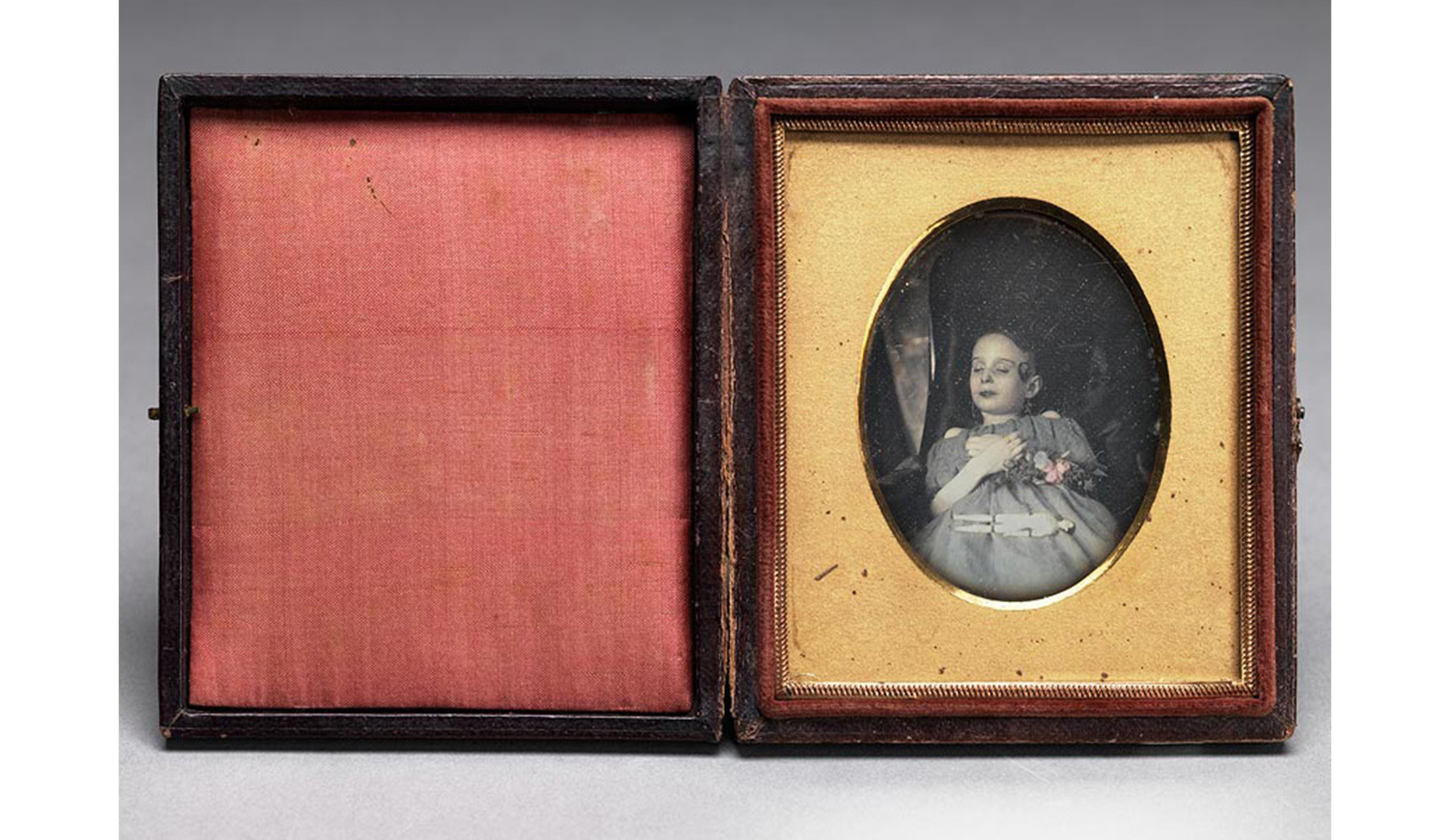brown leather tooled case with metal clasp, pink silk inside cover, plain gold mat around oval image of dead deceased girl; child with hands folded over chest in hand painted blue dress, hand colored flowers over her proper left arm and small jointed doll laying on her dress