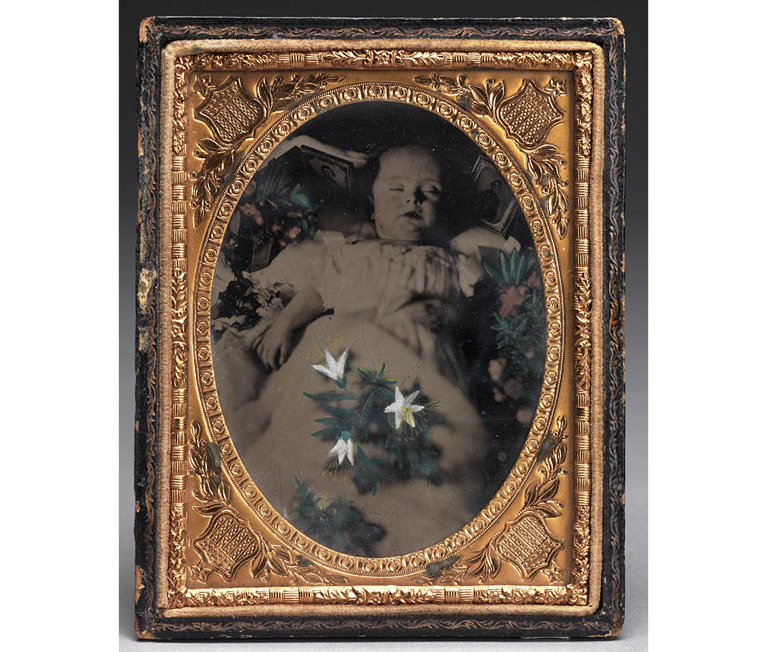 bottom half of black leather case with plain inset rectangle in center and two lines of gold braid tooling containing elaborate embossed mat (flowers and shields) with oval of deceased child, left hand bent over waist, right hand by side, partly covered in blanket, a small tintype on each side of her head, hand painted flowers on either side and over her blanket