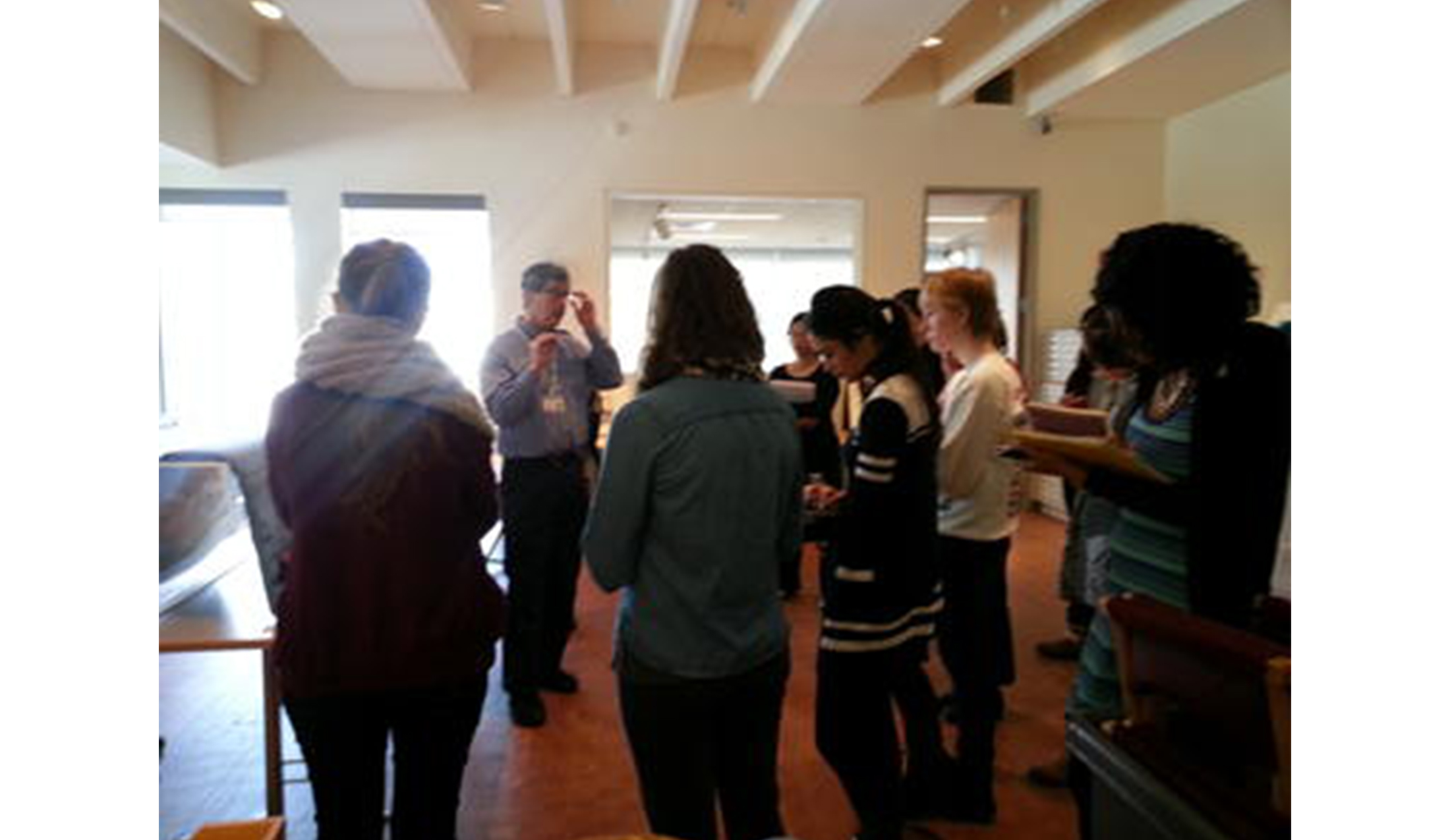 man stands in an art gallery in front of a circle of students, talking to them. the students are holding notebooks and taking notes