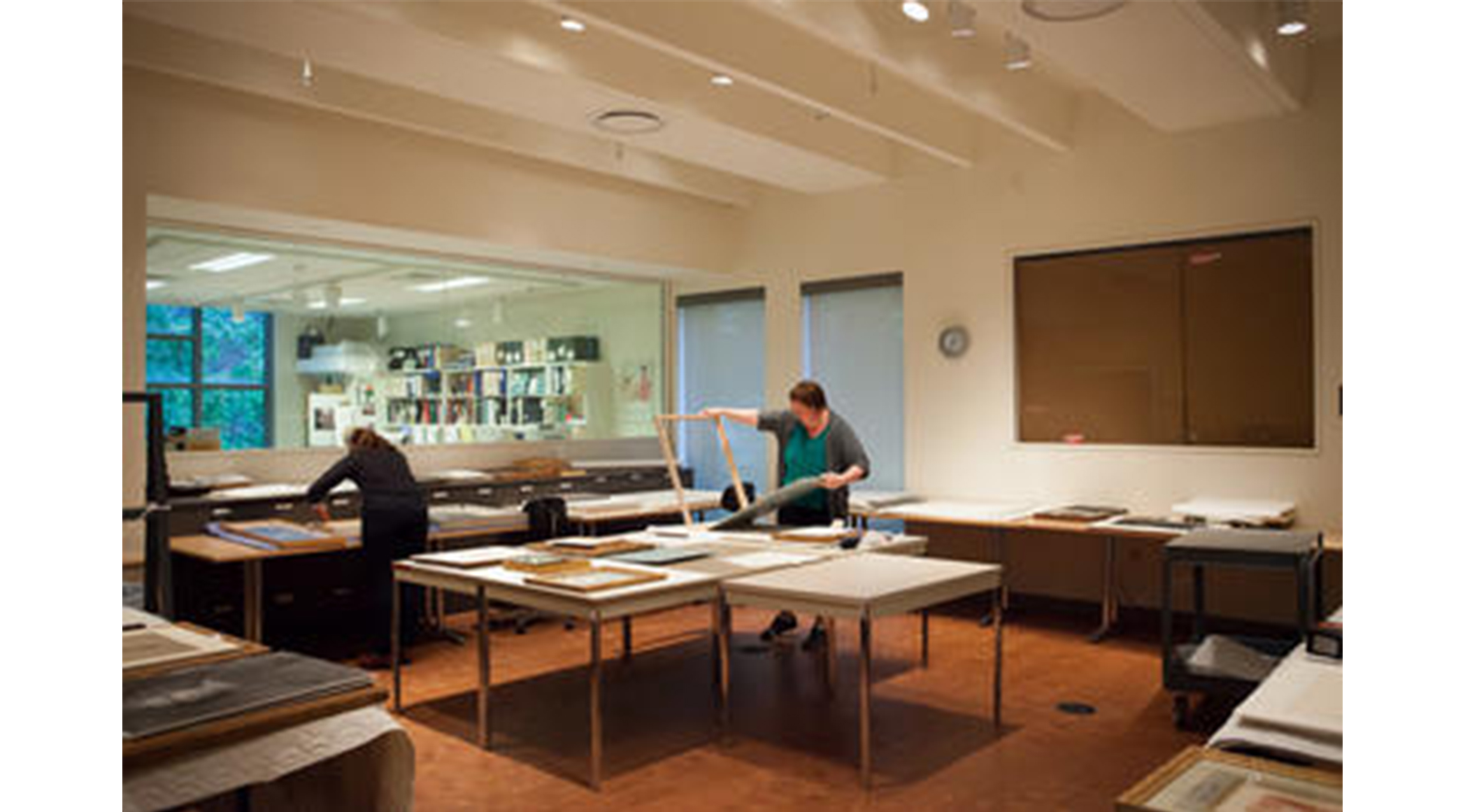 people standing in a large room with lots of tables. both are bent over a table handling framed artworks