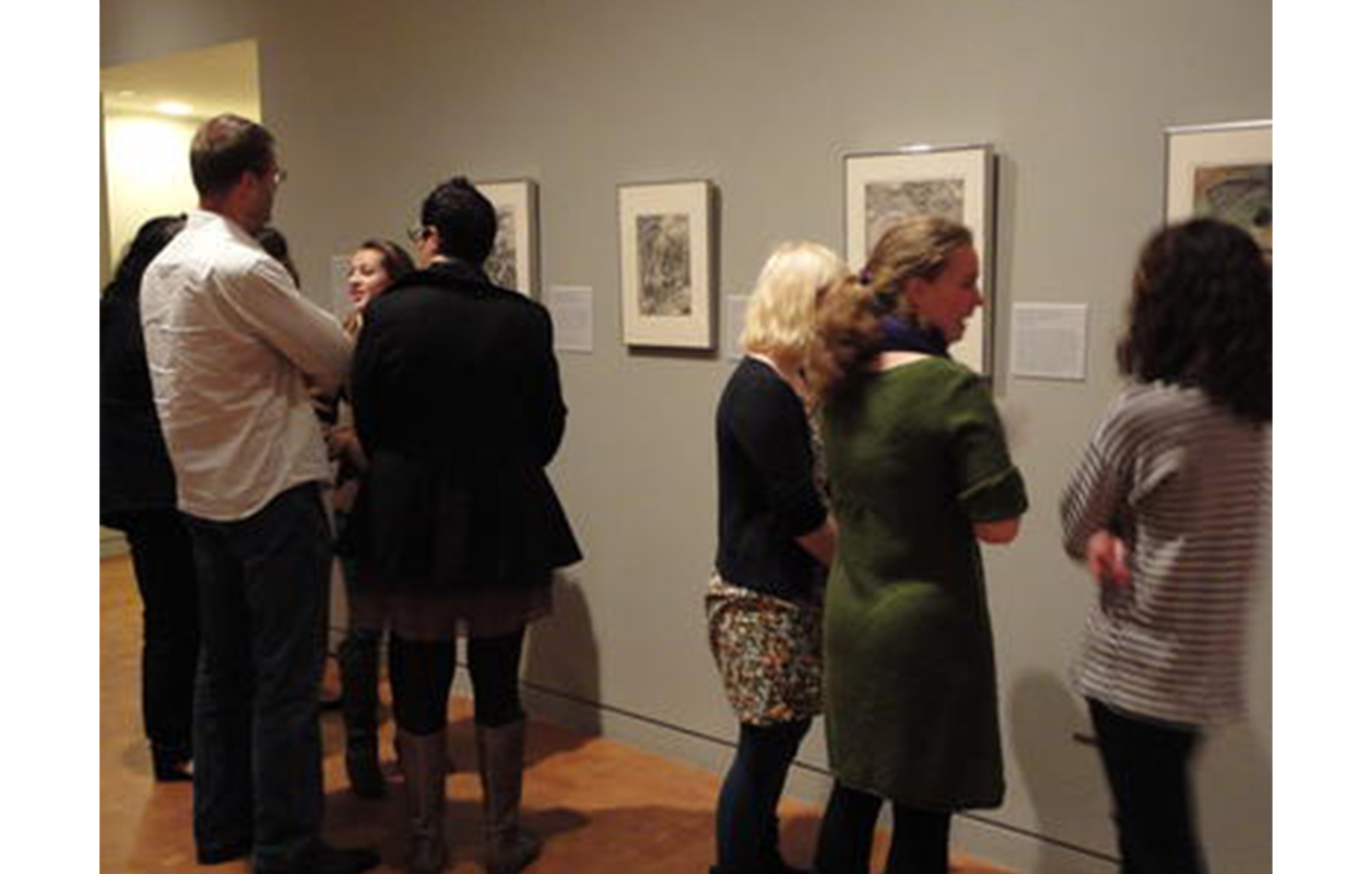 group of people dressed in formal clothing, looking at prints on a gray wall in an art gallery
