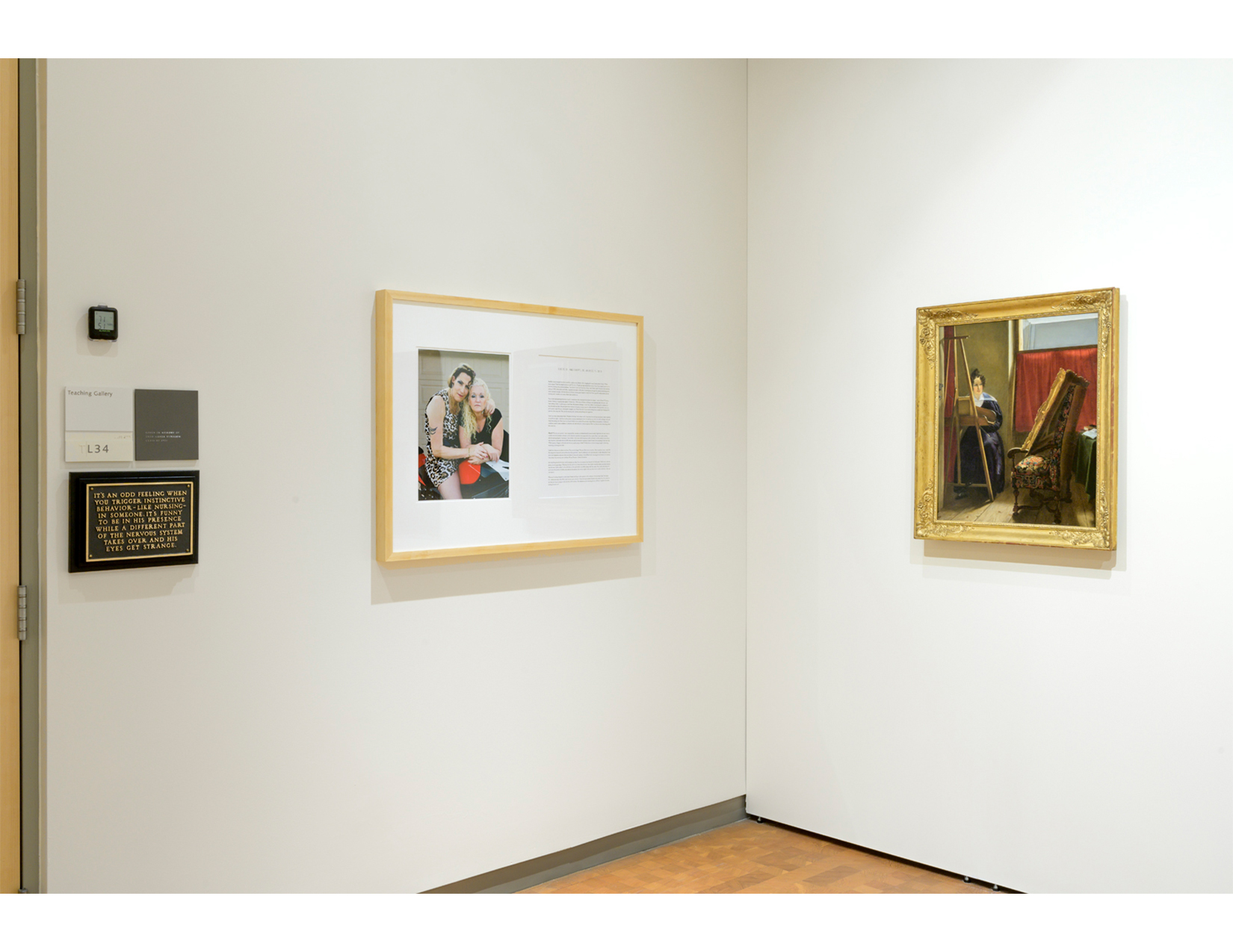 an art gallery with white walls, displaying three works of art. from left to right: a black plaque with gold words; an image of a couple embracing on a motorcycle, displayed next to a block of text; and a portrait of a woman painting 