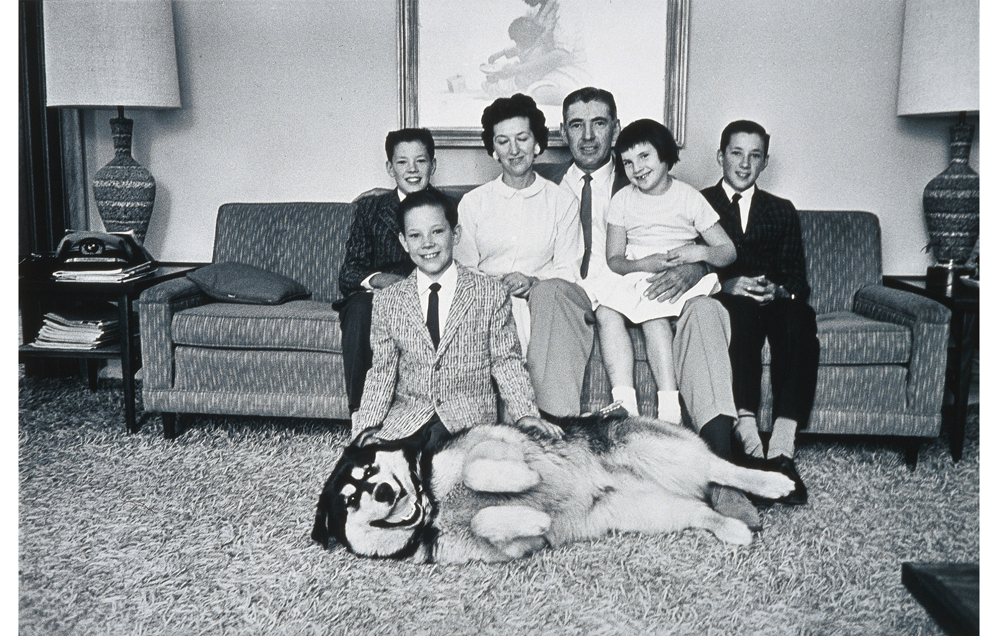 interior, husband wearing suit, wife wearing shirt and skirt, daughter wearing dress and three sons in jackets and ties on sofa with large German Shepherd lying across rug in front of them, end tables and lamps on either side of couch, painting on wall behind couch