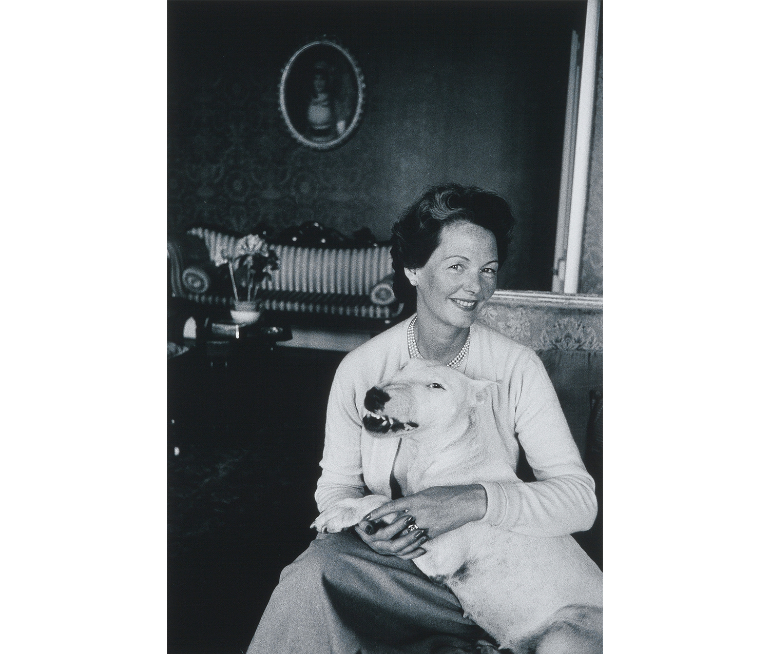 interior, woman in light sweater set and skirt, pearls, dark hair holding large white dog at front right, sofa, table, flowers, oval portrait visible in background