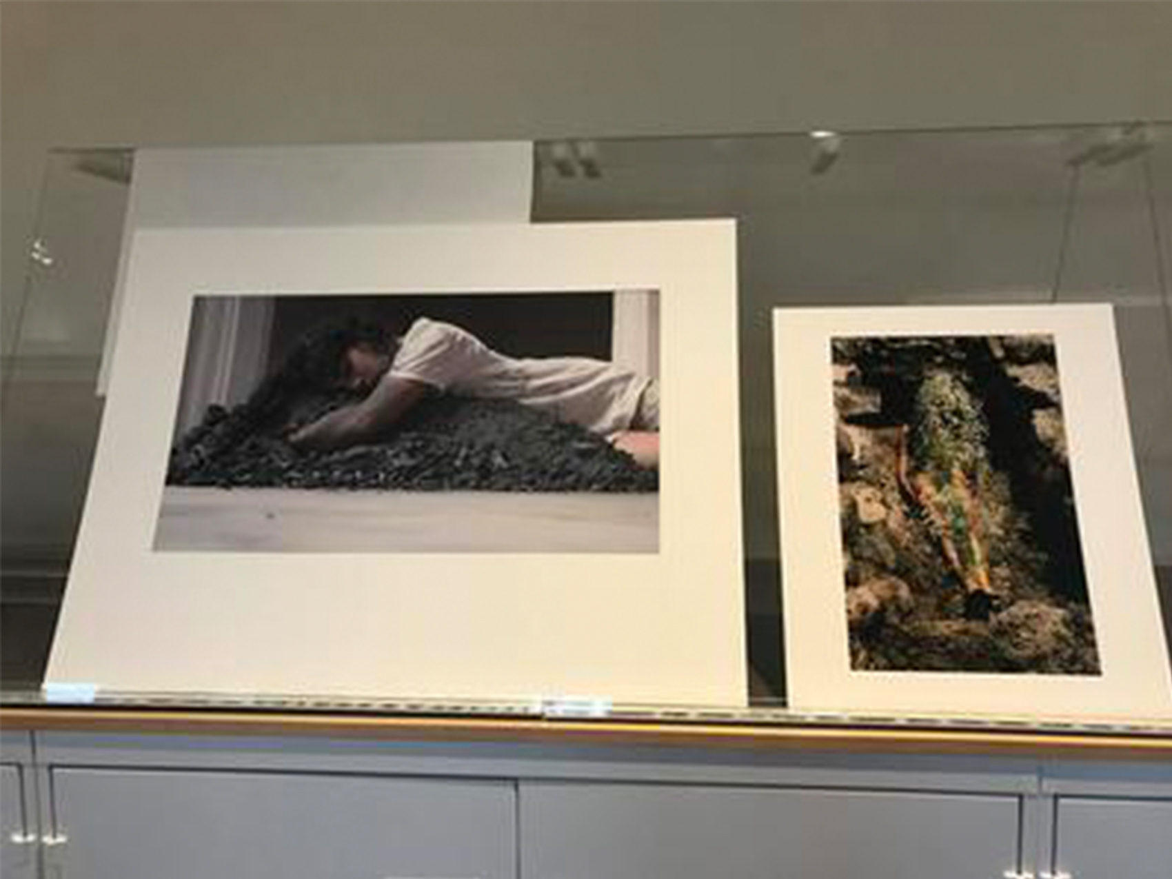 two prints propped up side by side, one of a woman lying in a pile of coal and one of a woman buried under leaves