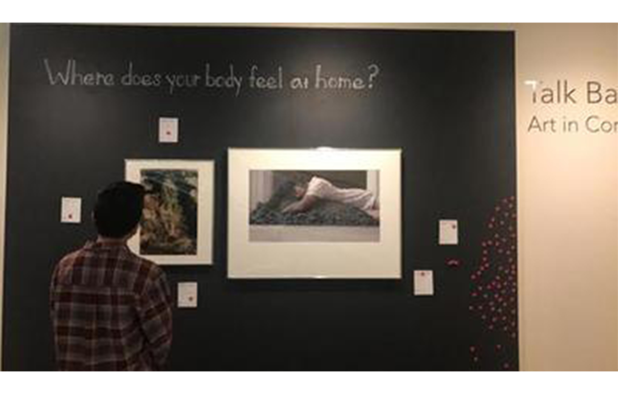 a person stands facing a black wall, on which two prints are displayed underneath the prompt, "where does your body feel at home?"