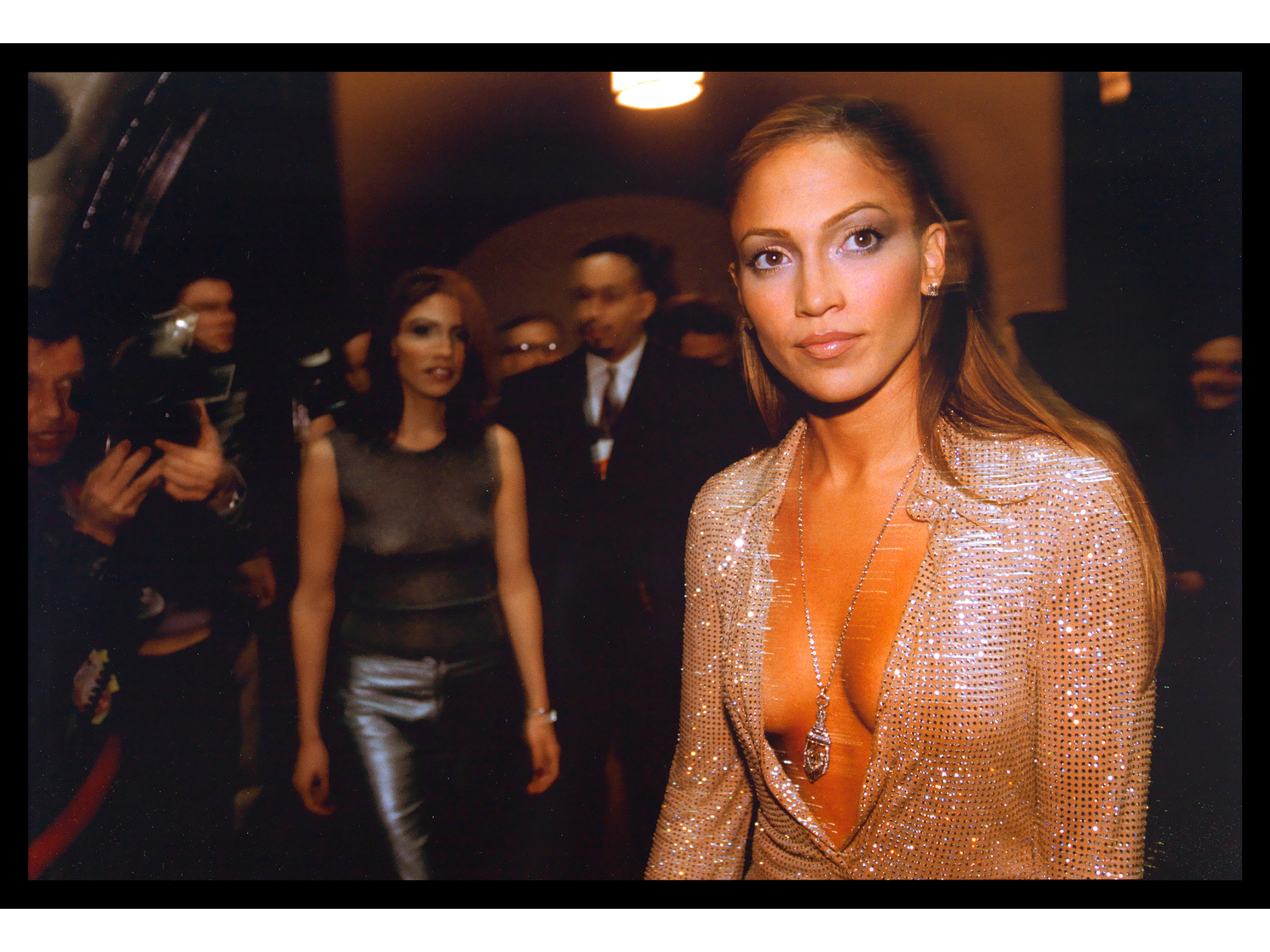 woman wearing a sparkly dress and silver necklace and looking up to her proper left. in the background, a blur of other people dressed in fancy clothing, one of whom is carrying a camera