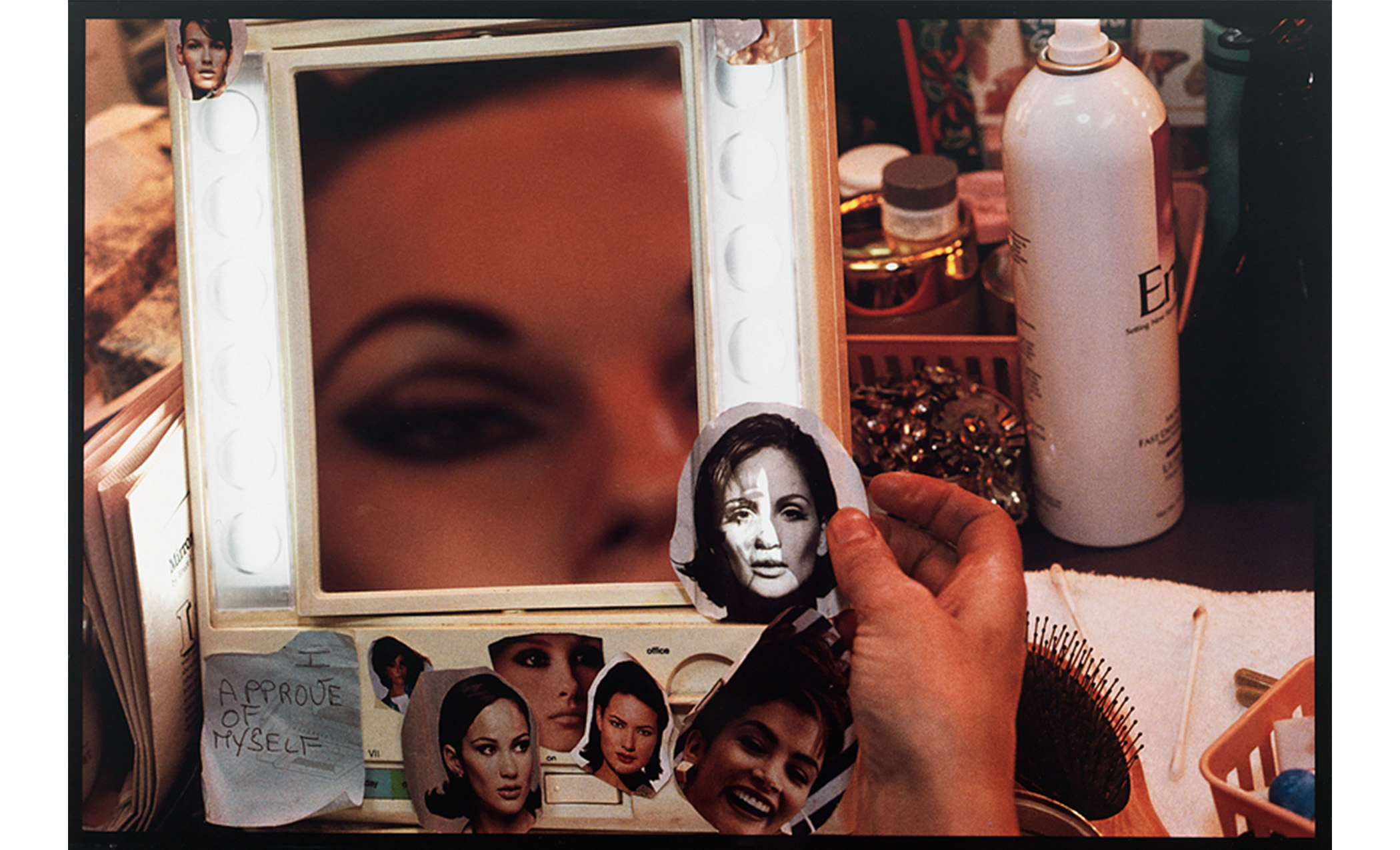 interior, close-up of a crowded dressing table with lighted mirror showing part of a woman's face as her hand holds the cut-out image of a model's head, other heads are attached to the mirror with a note saying "I / APPROVE / OF / MYSELF"