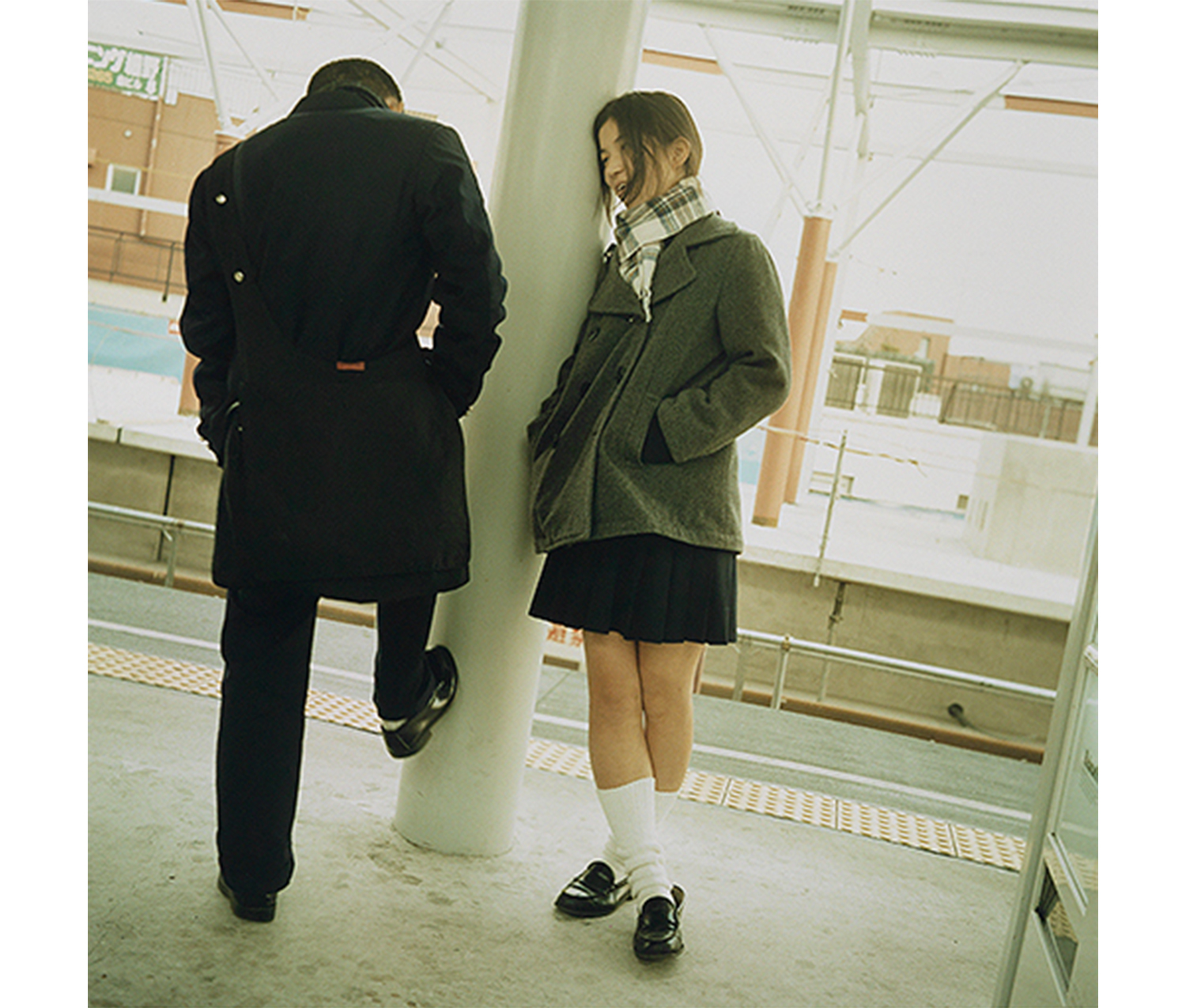 Two figures standing on a train platform. Girl on right wears plaid scarf, grey jacket, dark skirt, white knee socks and black shoes with her hands in her pockets leaning against a colum and leaning toward a boy on left wearing a dark jacket and pants seen from the back with his proper right foot braced against the column