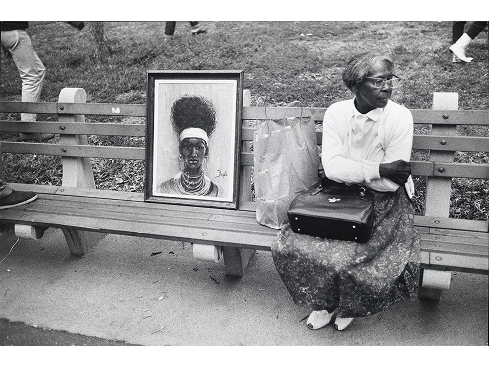 a black woman sitting on a bench wearing a blouse, skirt, and glasses with a purse on her lap and a bag on her right; also on the bench is a framed bust painting of a black person with extensive necklaces and large hoop earings, wide head band and high teased hair
