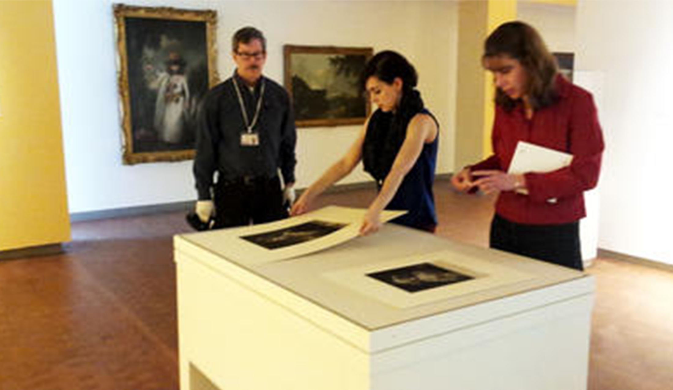 girl stands in an art gallery, placing a print onto the top of a cabinet. a man stands to her right and a woman stands to her left, both watching her work.