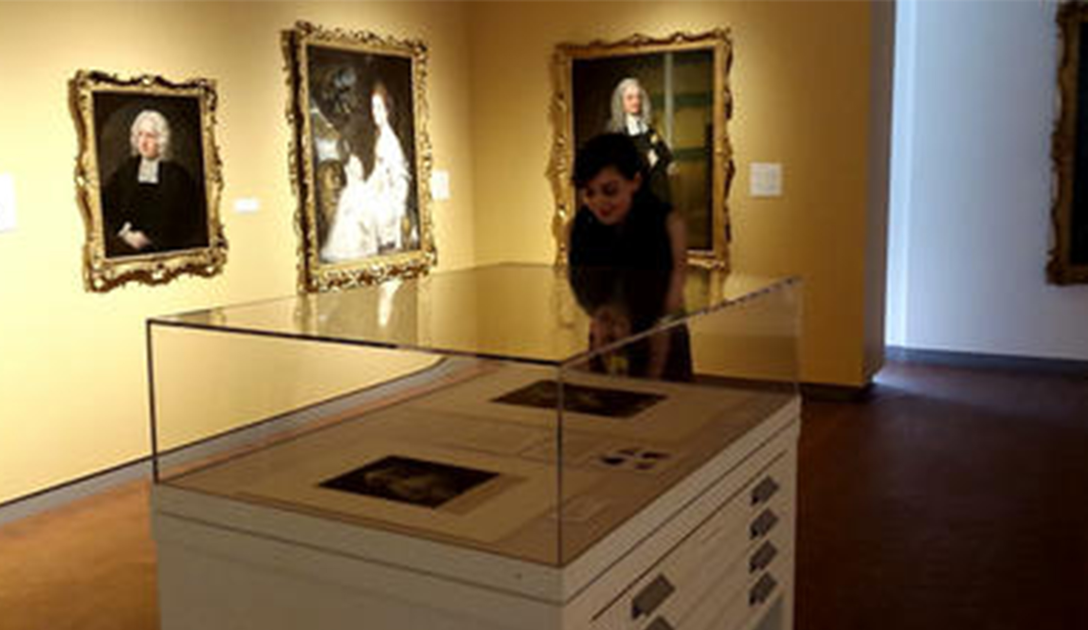 girl stands in an art gallery behind a cabinet with a glass display vitrine on top. 
