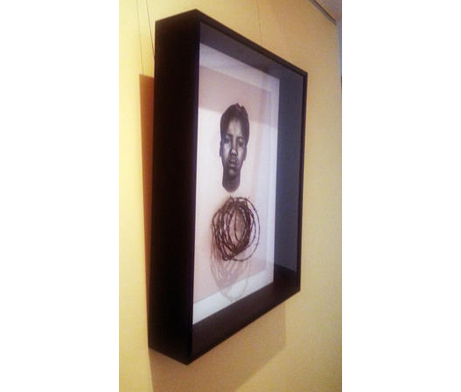 yellow wall with framed print hanging on it. the print shows the frontal head of a frowning African American woman with her hair in an up do over a circlet of real barbed wire encased in a black box