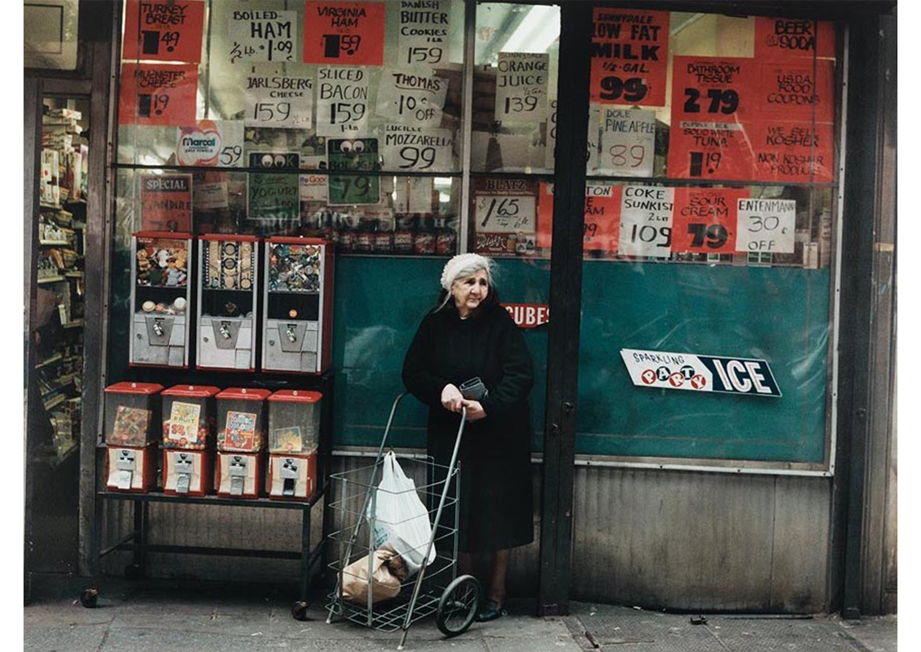 older woman in dark coat, white hat and pull cart standing in front of a grocery store window plastered with signs with group of gum ball machines to her proper left