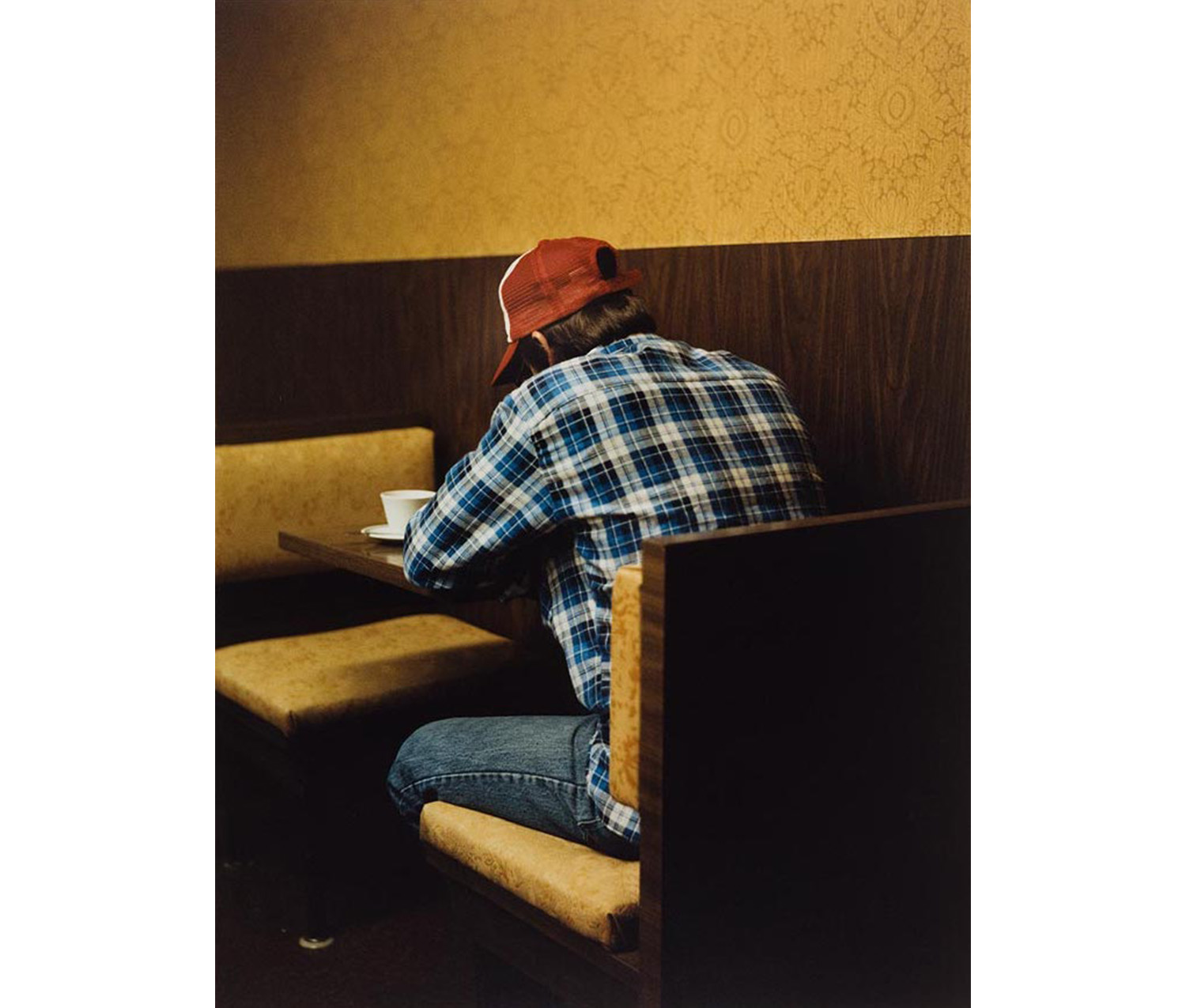 interior, back of a man with red baseball hat, blue plaid shirt and jeans seated at a booth made of wood grain laminate and gold cloth with a cup of coffee on the table before him