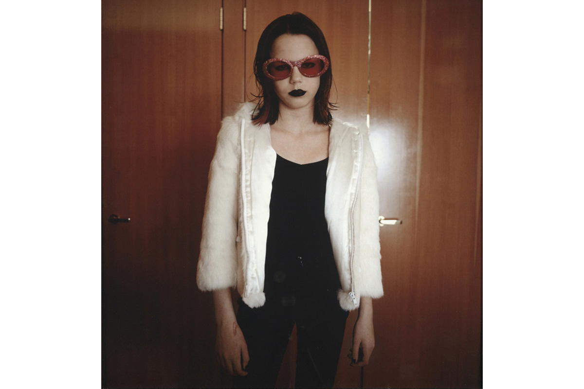 "figure facing forward with dark hair highlighted with fuschia wearing pink sparkly sunglasses and black lipstick and wearing black top and pants and a furry white jacket"