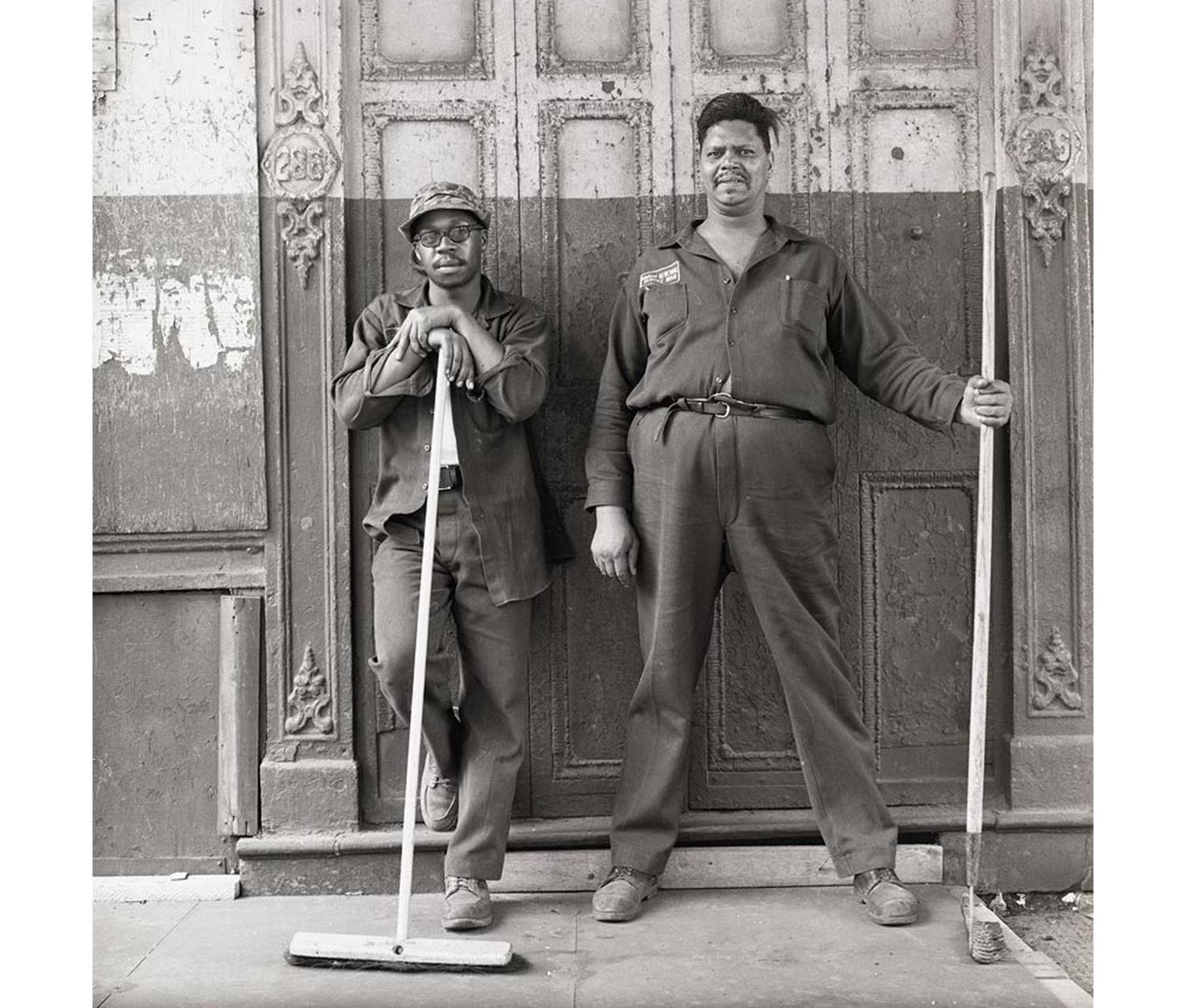 two men in work clothes, one holding broom, the other a rake, standing in front of old painted doors of closed building, each looking seriously into the camera