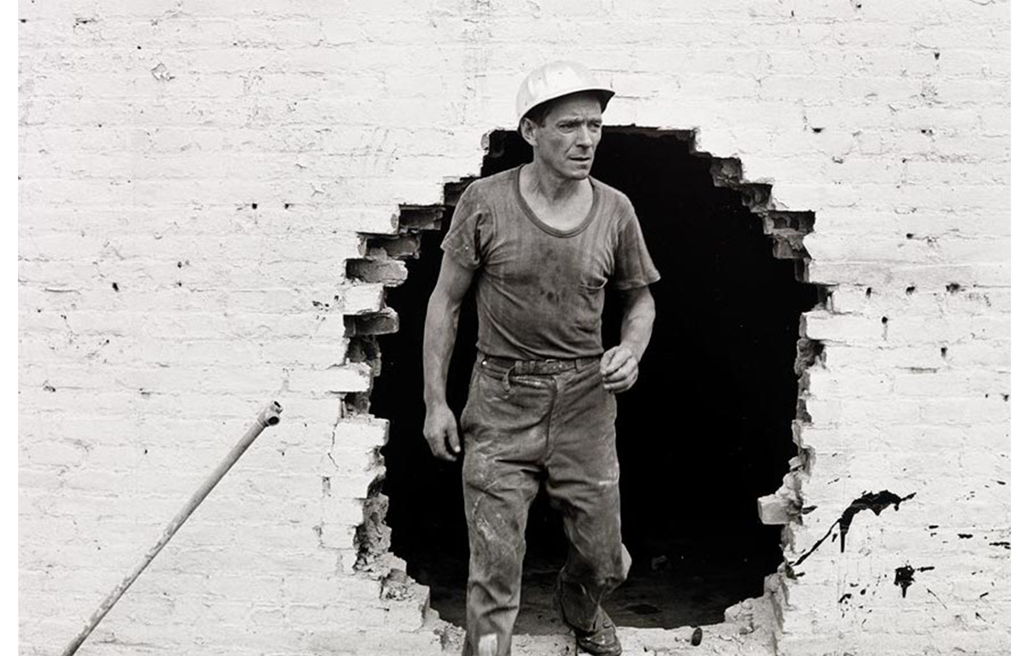 man in dirty T shirt and jeans with hard hat coming out of a large hole in a white painted brick wall, a diagonal pipe sticking up at the lower left