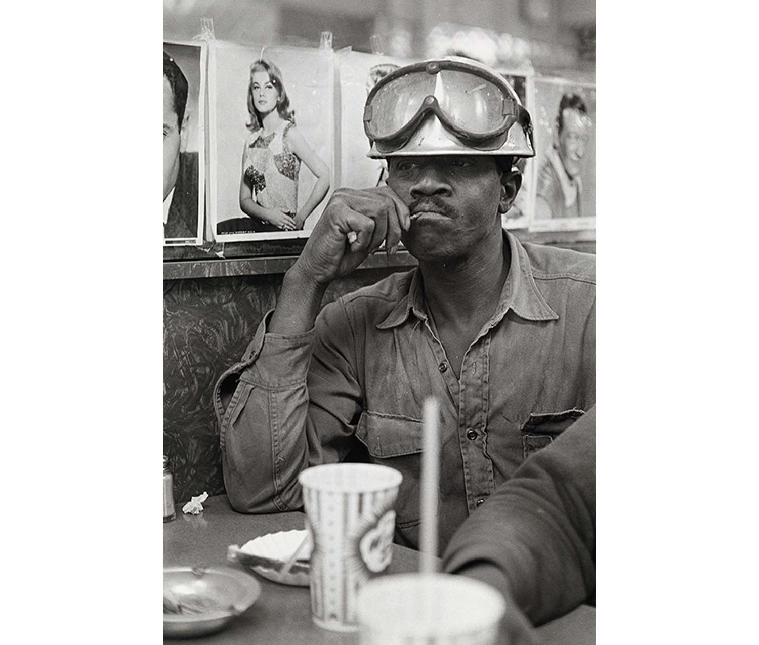 man in work clothes with hard hat and protective glasses on top of hat, seated at table with celebrity portrait photographs in background