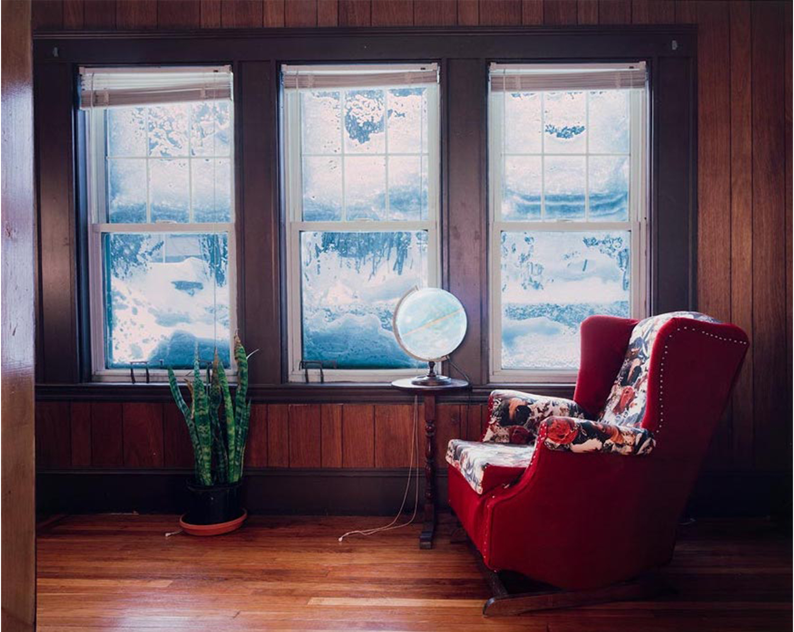 interior, wood paneled room, wood floor, succulent in pot on floor at left, globe with interior light on small round table near padded rocker with red upholstery on sides and back and animal print upholstery on cushion, arms and interior back horizontal to three windows that heavily frosted over barely showing snowy landscape outside