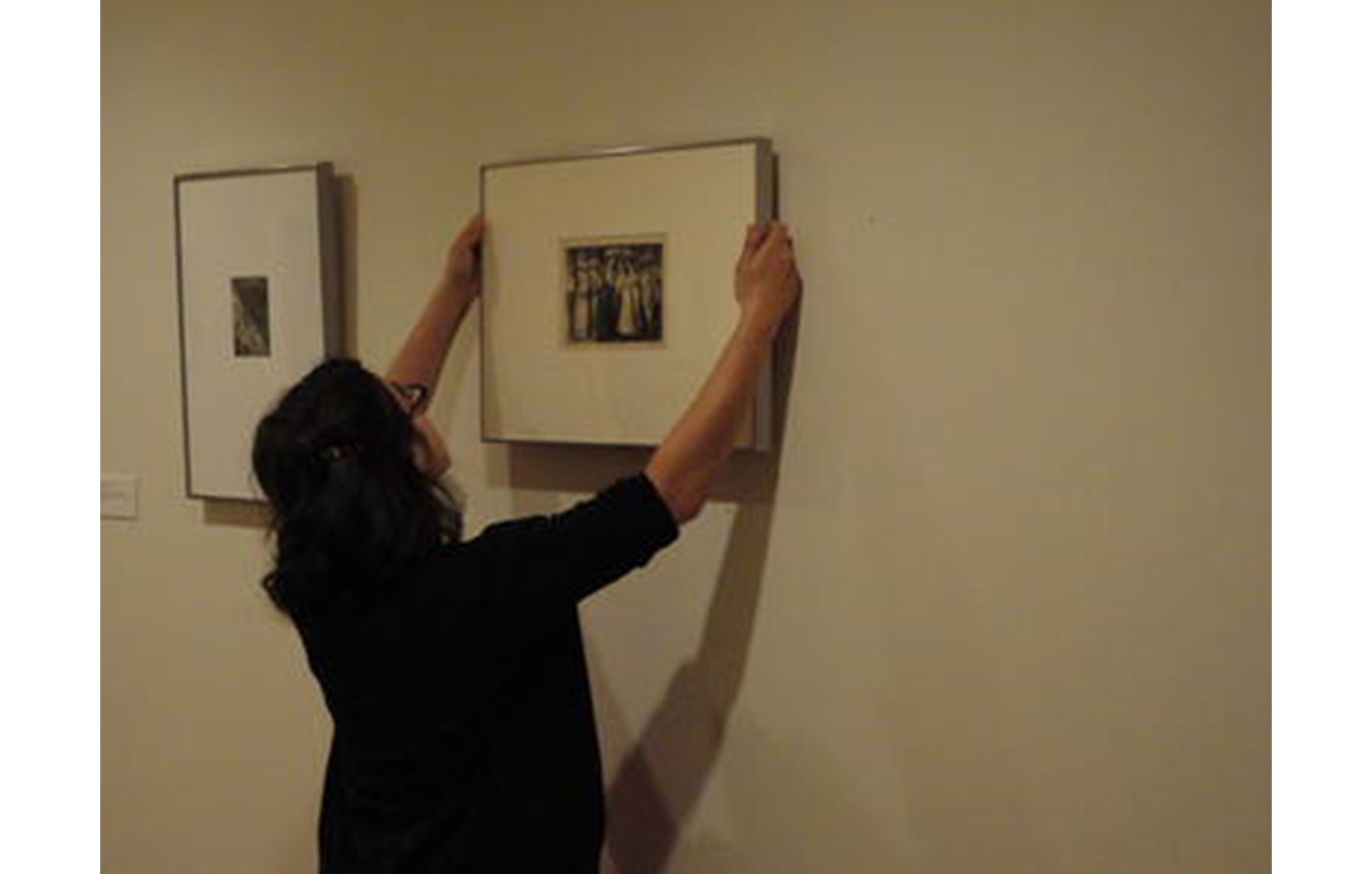 woman with dark hair and classes stands in an art gallery, hanging a print on the wall to the right of another print that is already hung on the wall