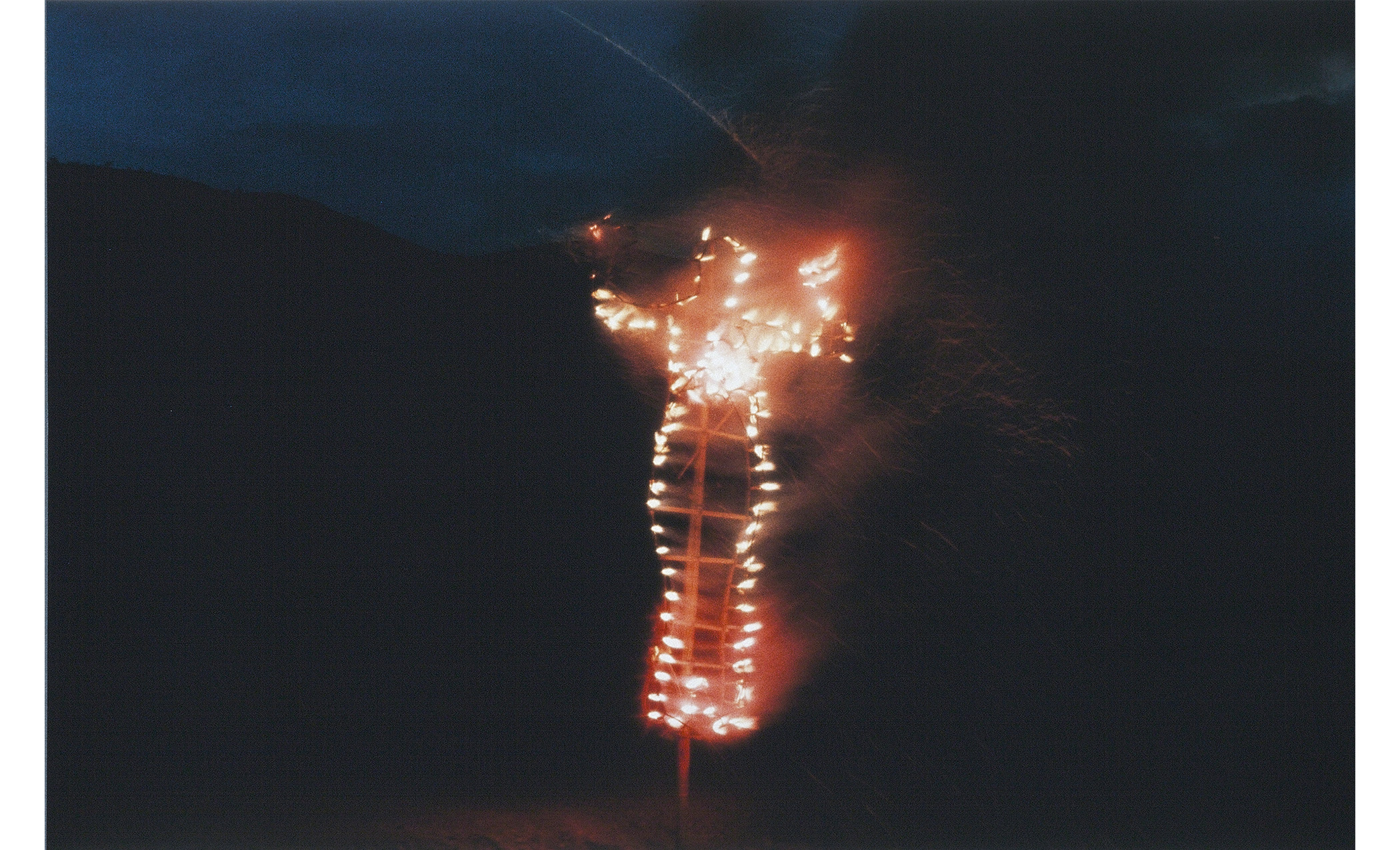 night, dark sky, low dark hills in background, burning bamboo frame in shape of woman with arms upraised on a pole in the foreground