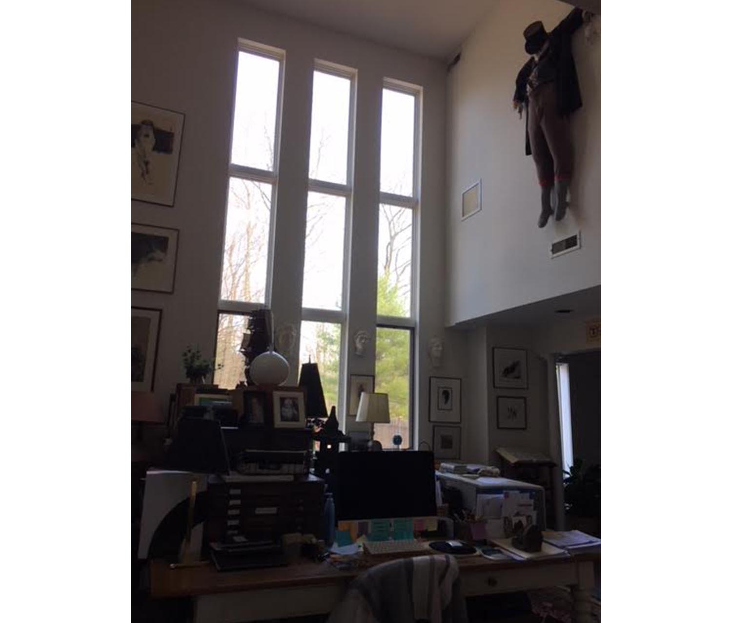 an artist's studio, with a desk containing picture frames, a printer, and a computer. in the background, a sculpture of a body hangs from the ceiling and many trees can be seen through three floor-to-ceiling windows 
