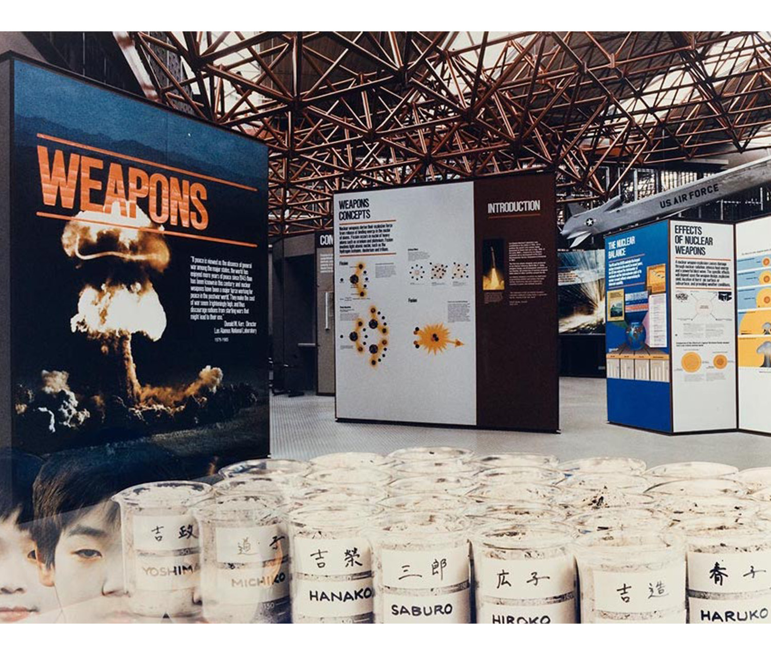 interior, large room with exhibition panels, vials with names on them across the front and overlaid image of Japanese children at lower left corner