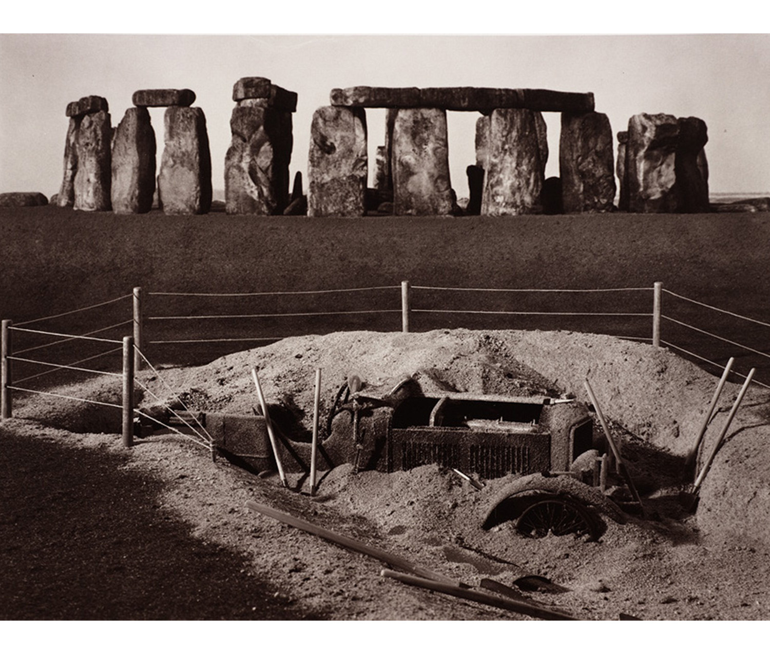 outdoor, Stonehenge visible in background, hole surrounded by wood and rope fence with shovels lying around it contains partly visible car