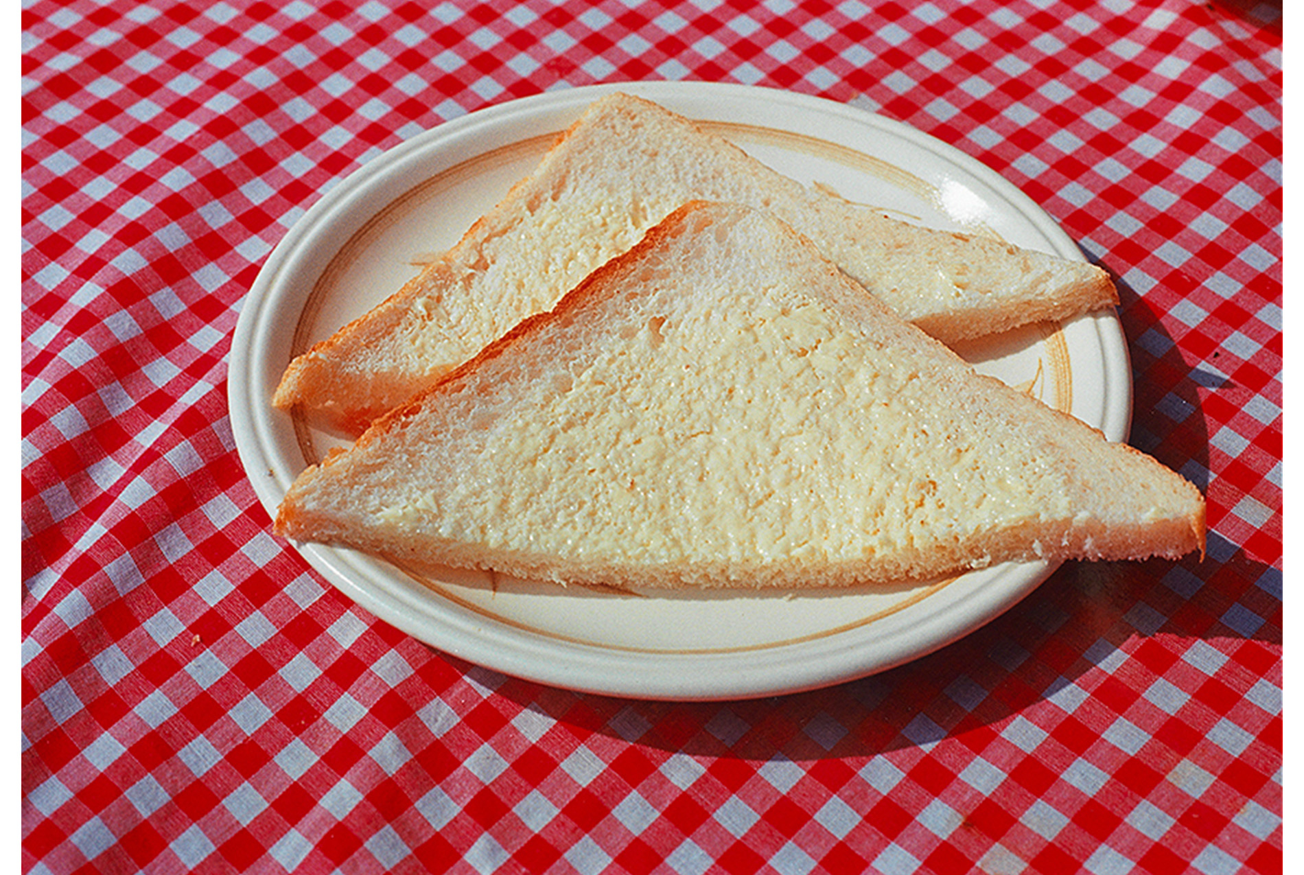 close-up of a table with a red and white checked cloth, white plate with gold band around inner edge holding two slices of thin white bread with spread with butter