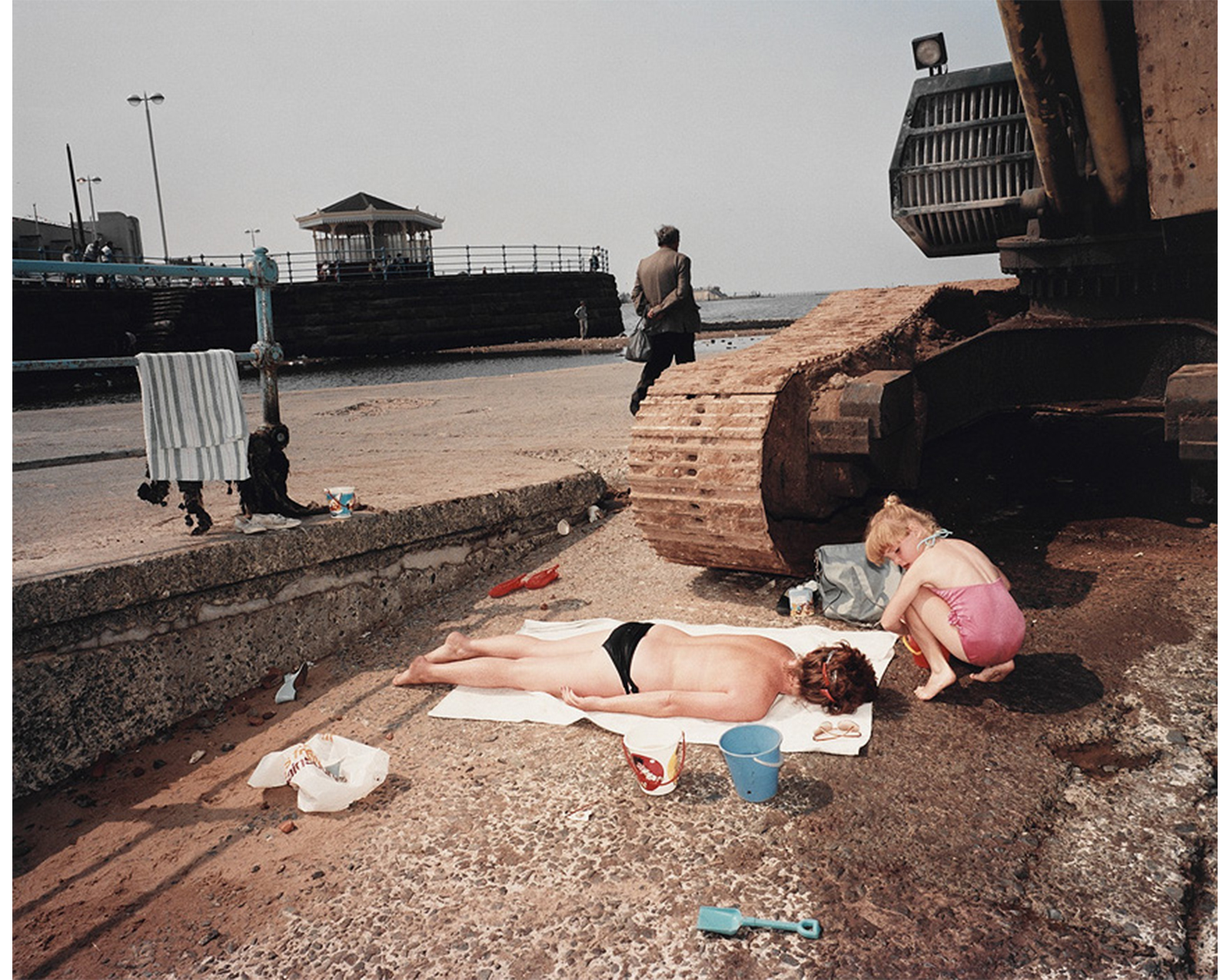 beach, walkway visible in back left with lights, small building and people, man wearing jacket walking toward water in center holding a bag, rusty tank at left edge with woman on her stomach on a white blanket sunbathing topless, a blond child in pink bathing suit crouching beside her holding a sand pail