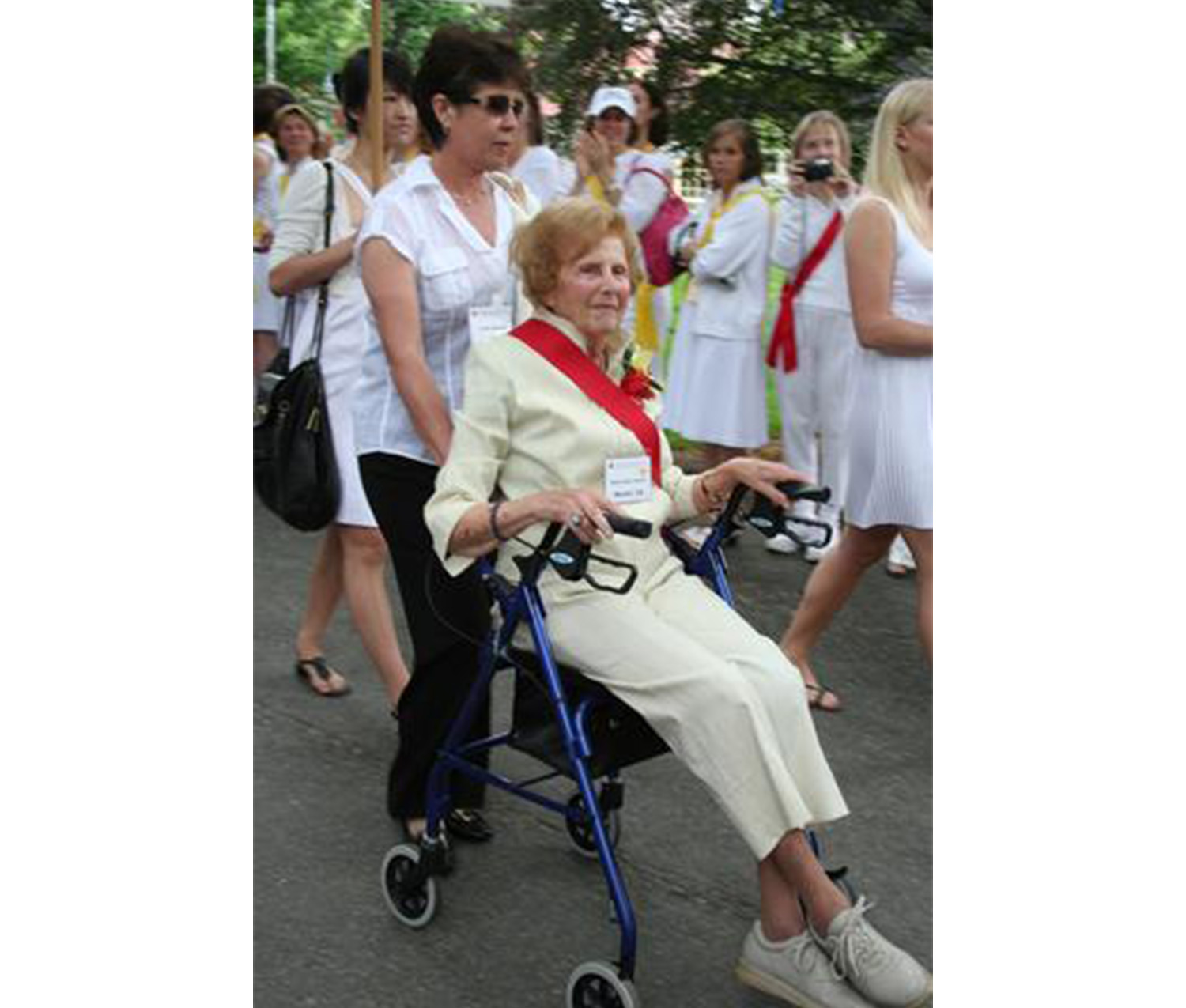 woman wearing white dress and red sash sits in a wheelchair; another woman wearing white is pushing her, and several other women in white walk behind her