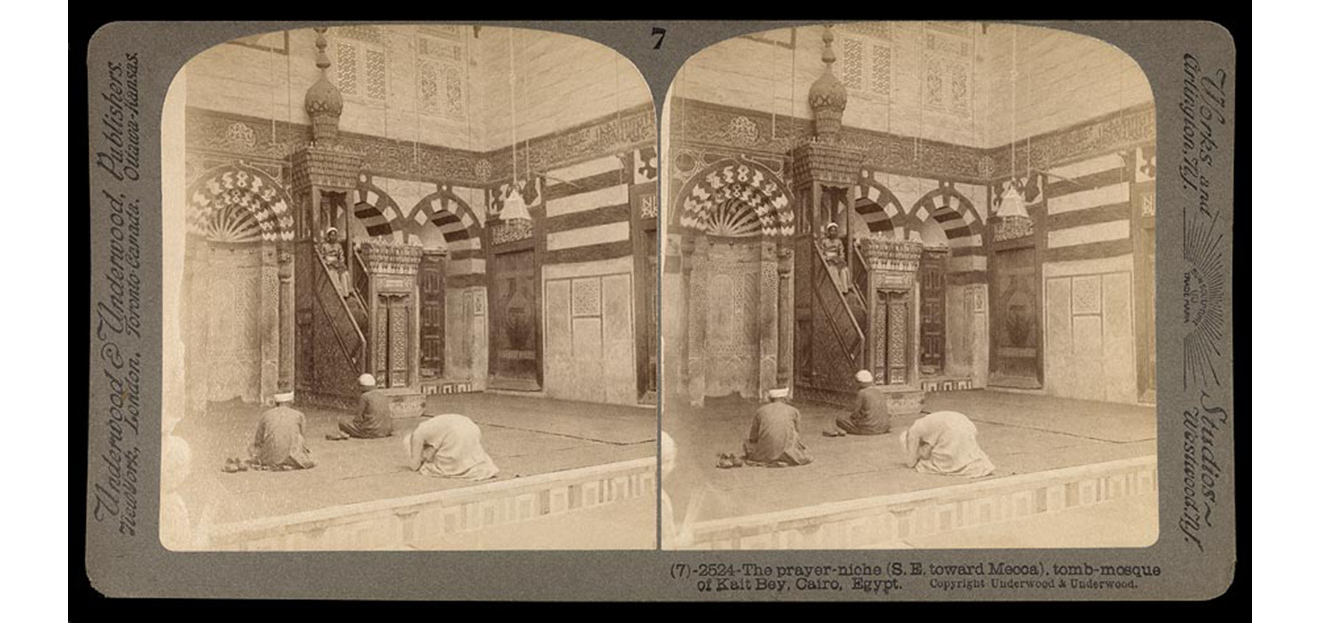 elaborate interior with three men, two sitting and one kneeling forward, all in robes and turbans praying as another man sits on top step of an elaborate covered staircase