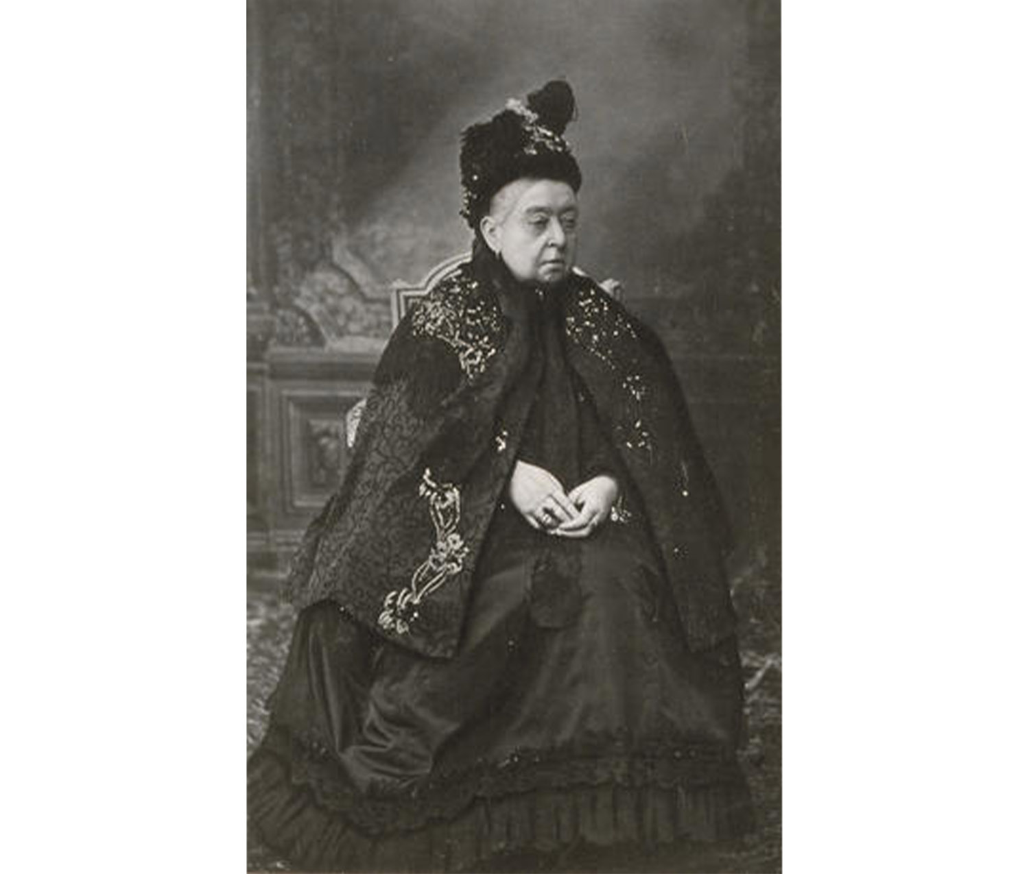 woman wearing long black dress and cape sits on a throne, looking to her left