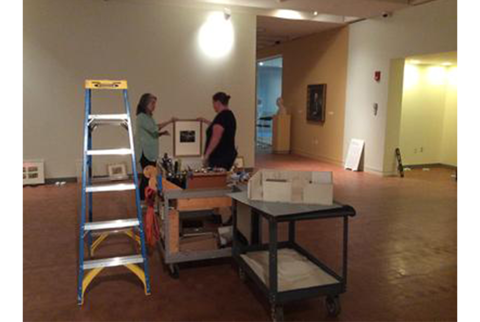 two woman holding a framed print in an art gallery, preparing to hang it up. in front of them sit a blue ladder and a white cabinet
