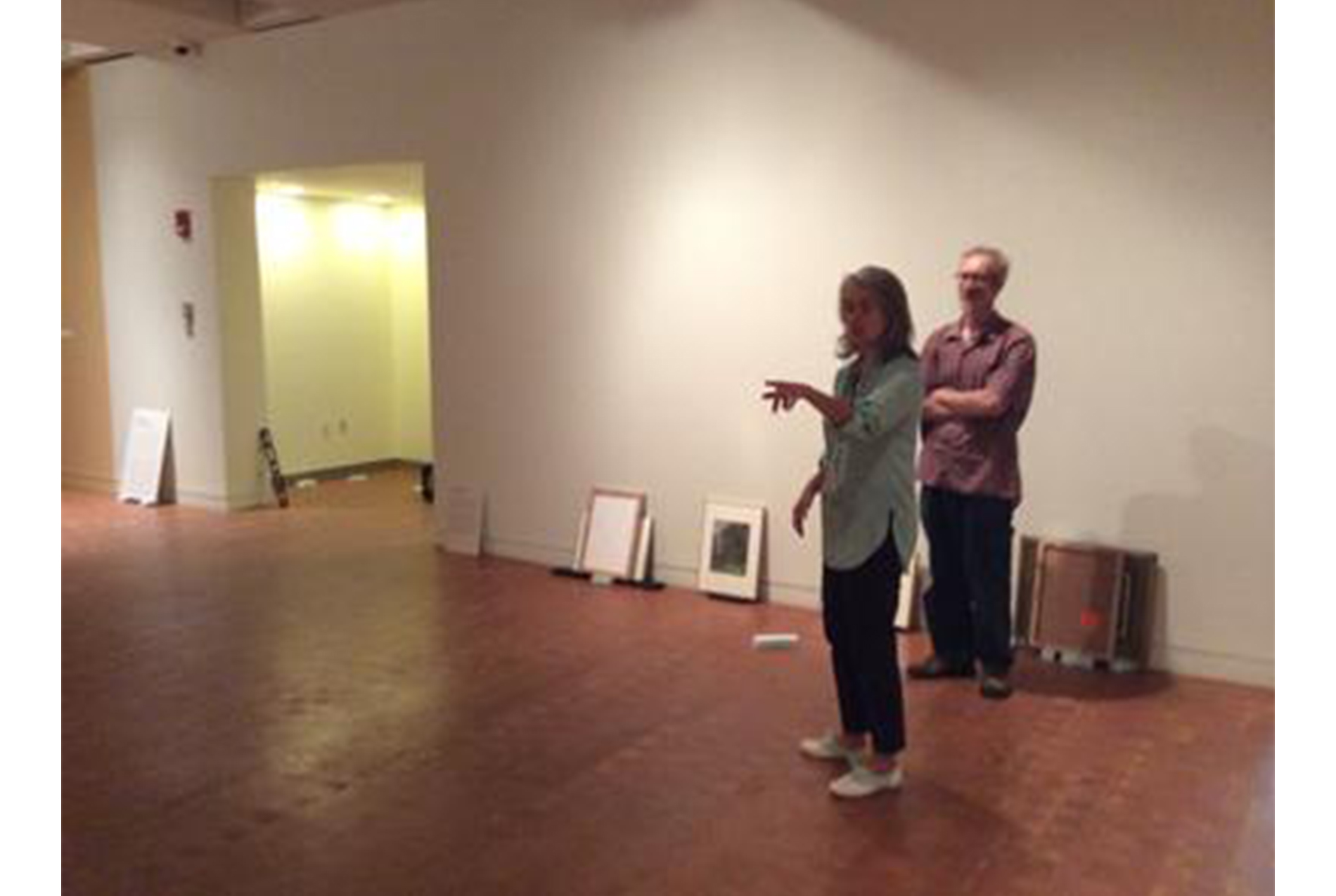 woman stands in an empty art gallery, pointing to her right. man stands behind her with his arms crossed
