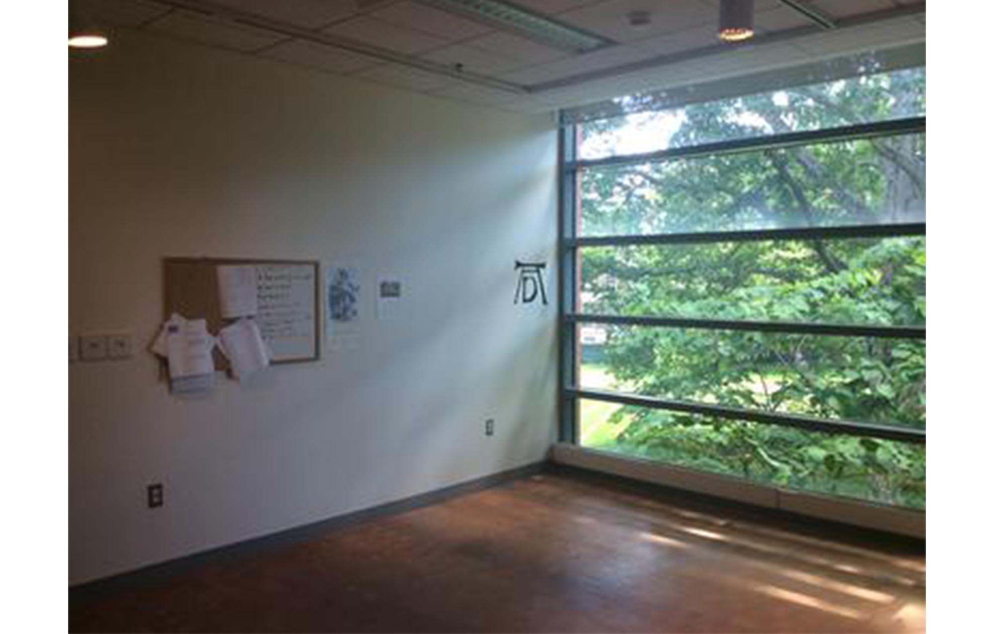 empty room with wood floors. back left wall is plain white and has a whiteboard and cork board on it. back right wall has floor-to-ceiling windows showing green trees outside