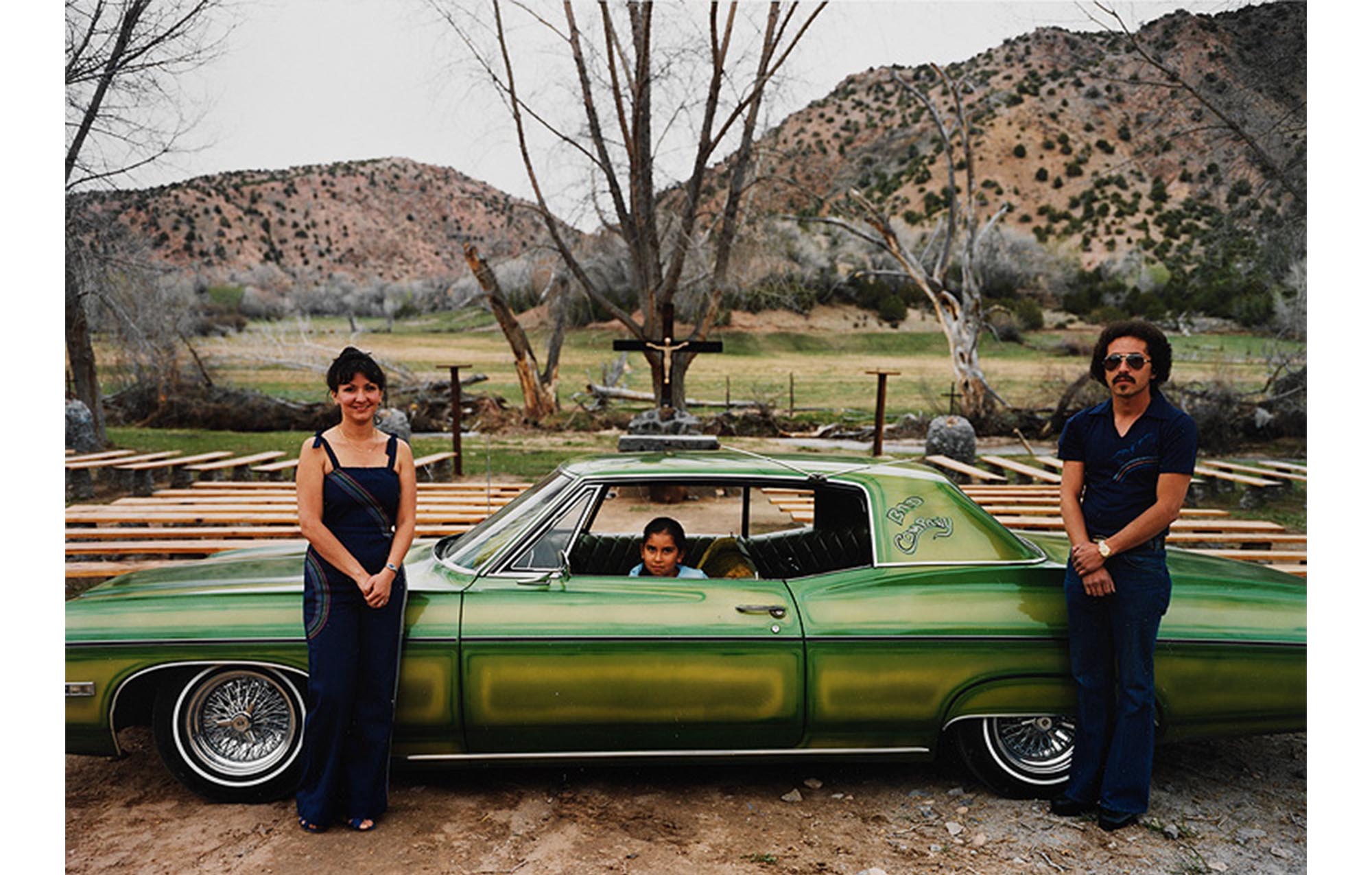 rural roadside, hills in distance with scrub growth, roadside shrine with crucifix in center and benches placed around it, in foreground a green sedan with lighter green painted panels, small child seated in driver's seat, young woman to left standing near hood in blue denim slacks and top, young man with mustache in same patterned denim outfit standing near trunk
