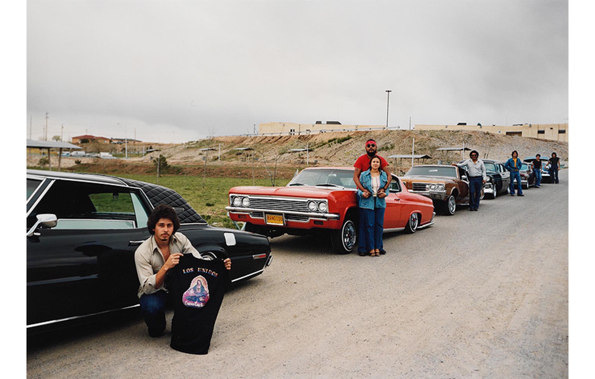 desert road, large low building in background, row of cars parked by the side of a dirt road with their owners standing near them, the owner of the first car in line squatting down and holding a black T-shirt with the Virgin and Los Unidos printed on it
