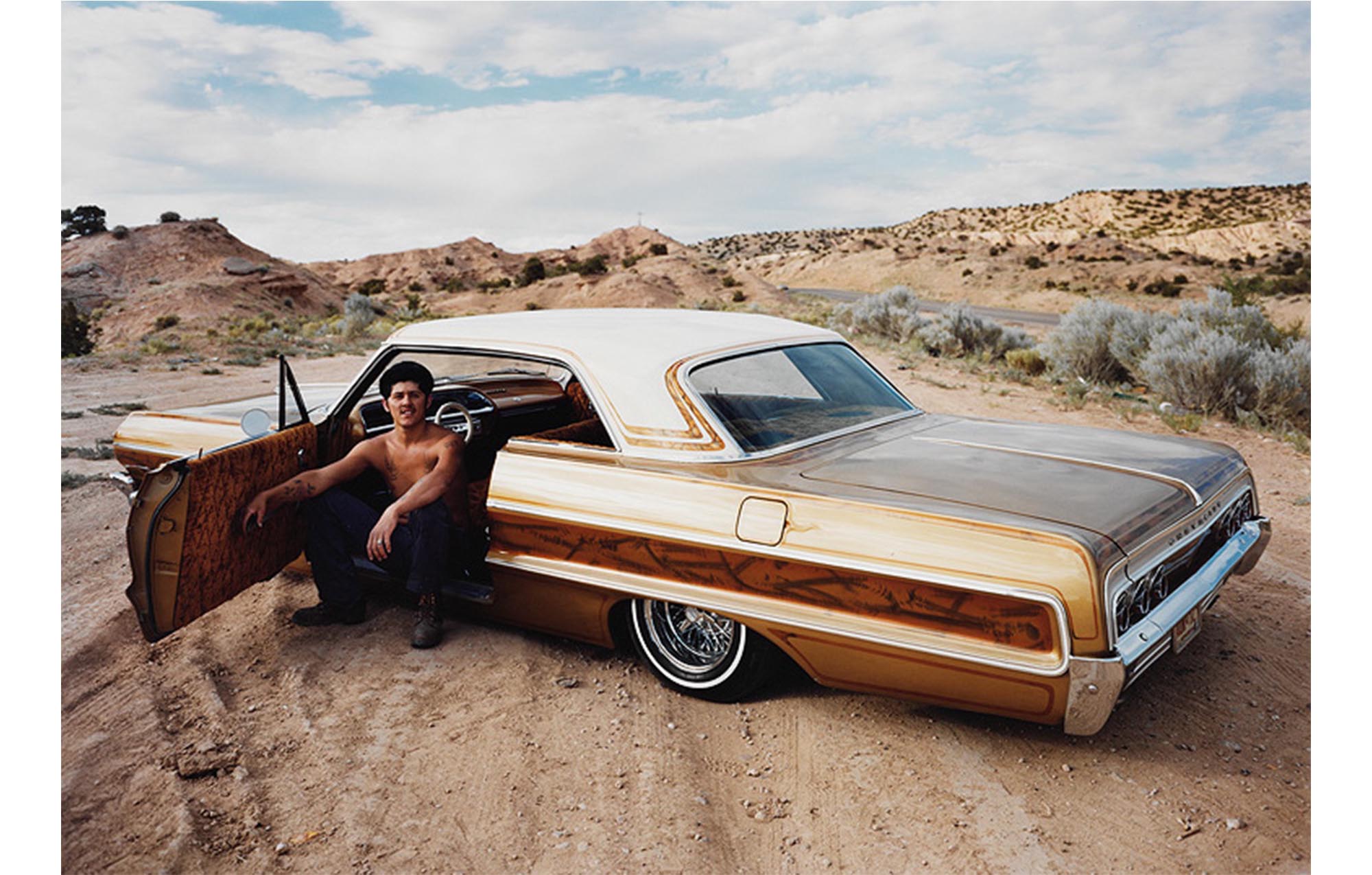 desert, low hills and scrubby growth, gold sedan with white top and bronze design in foreground, bare chested young man in blue jeans sitting in driver's seat