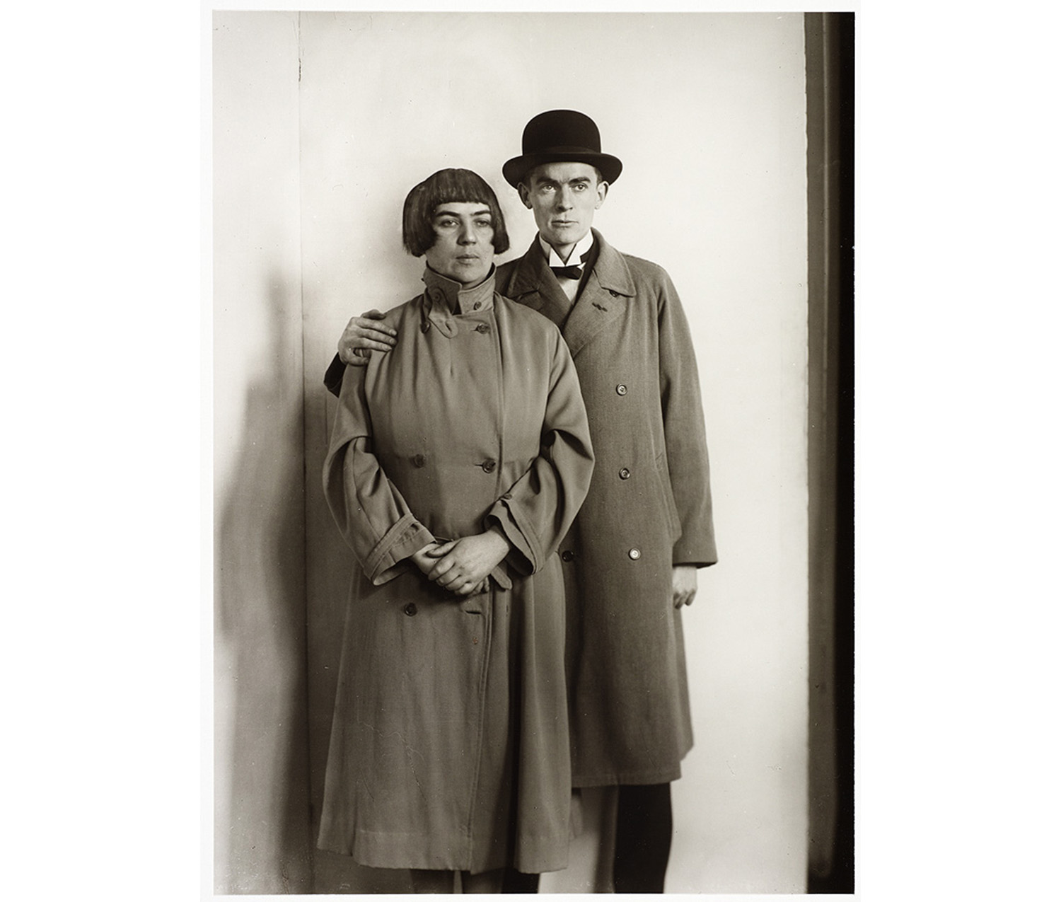 portrait of standing man and woman near a corner of a white wall; woman has cropped hair, a serious expression and wears a completely buttoned coat with the collar up, hands joined in front of her; man has a bowler hat, serious expression, proper left hand on her shoulder, wearing a buttoned overcoat, stand-up collar and black tie