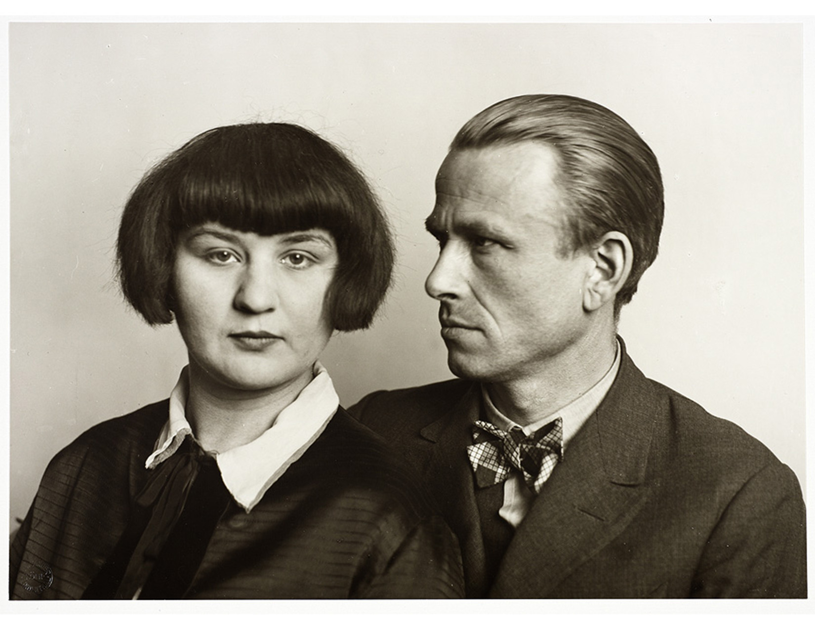 bust portrait of a couple against plain backdrop; woman has a dark blouse with white collar, she has cropped hair and a serious expression looks out at the camera; the man seated overlapping with her has a suit, light shirt and printed bow tie, he has slicked back hair, a serious expression and is looking at her